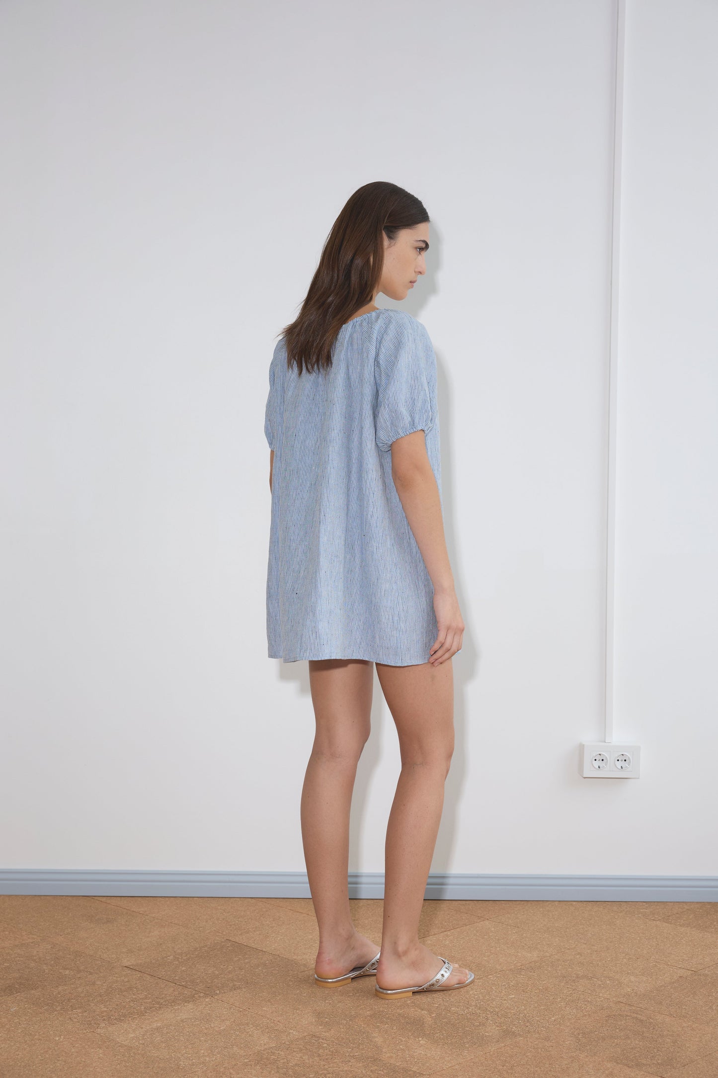 A woman with long brown hair stands sideways in a minimal room, wearing the Smock Dress - Blue Stripe and sandals. The wall behind her is white with two electrical outlets near the floor.