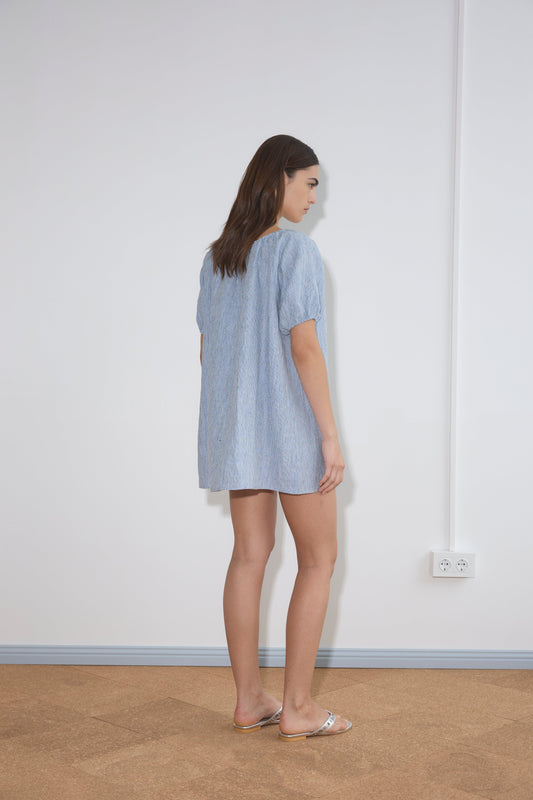 A woman with long brown hair stands sideways in a minimal room, wearing the Smock Dress - Blue Stripe and sandals. The wall behind her is white with two electrical outlets near the floor.