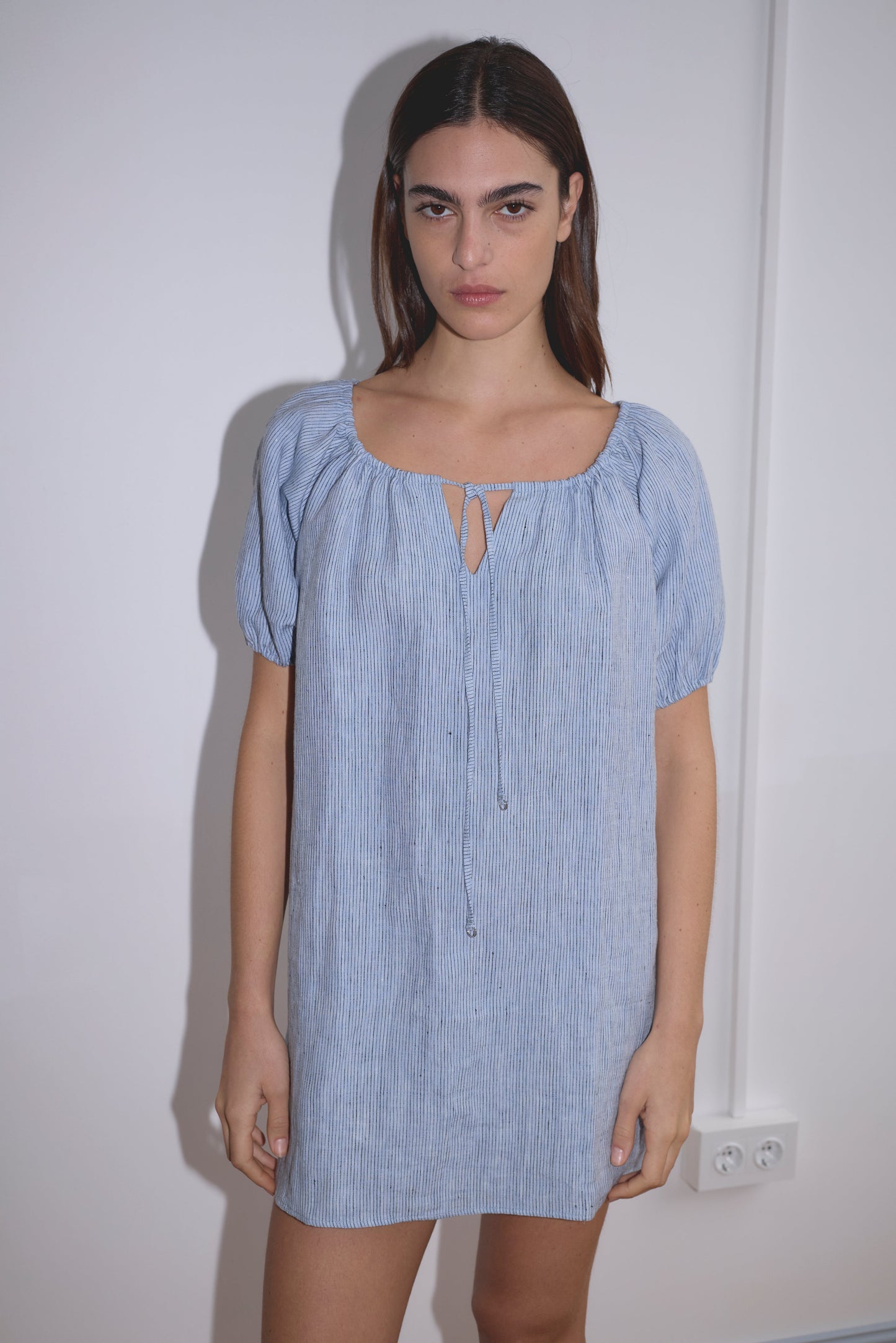 A woman with long brown hair stands against a white wall, wearing the Smock Dress - Blue Stripe featuring subtle vertical stripes and a keyhole neckline. She looks directly at the camera with a neutral expression.