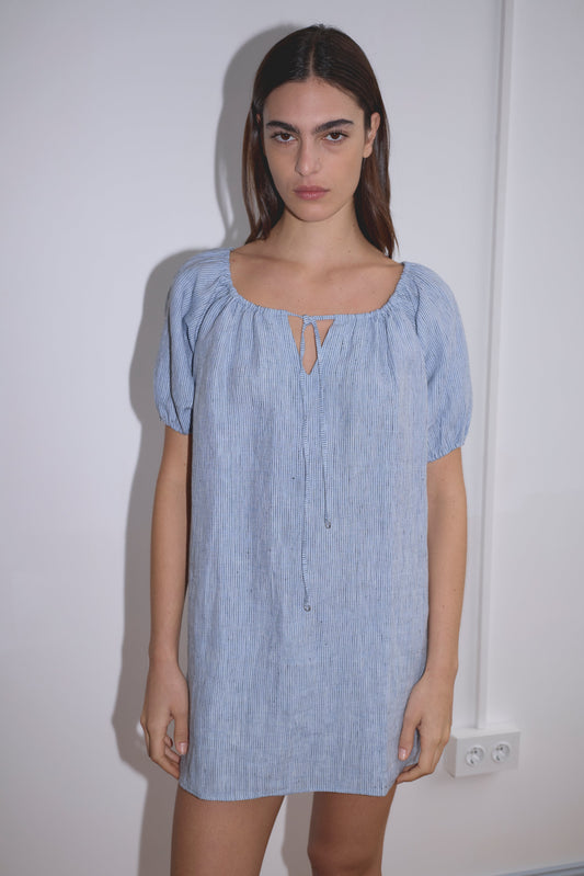 A woman with long brown hair stands against a white wall, wearing the Smock Dress - Blue Stripe featuring subtle vertical stripes and a keyhole neckline. She looks directly at the camera with a neutral expression.