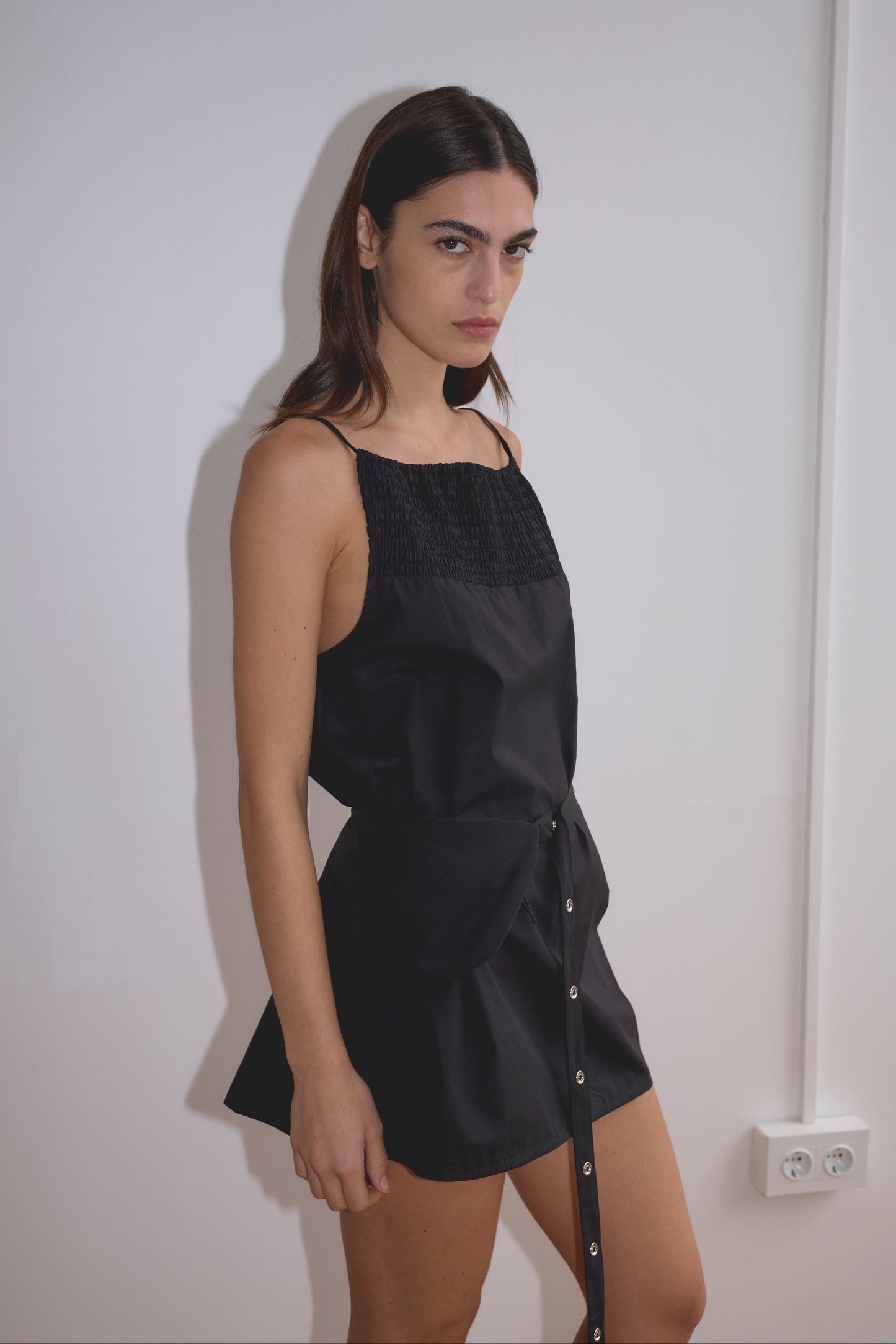 A woman with long dark hair stands against a plain white wall, wearing the Shirring Mini Dress - Black by Deiji Studios. She has a serious expression, and a white electrical outlet is visible on the wall nearby.