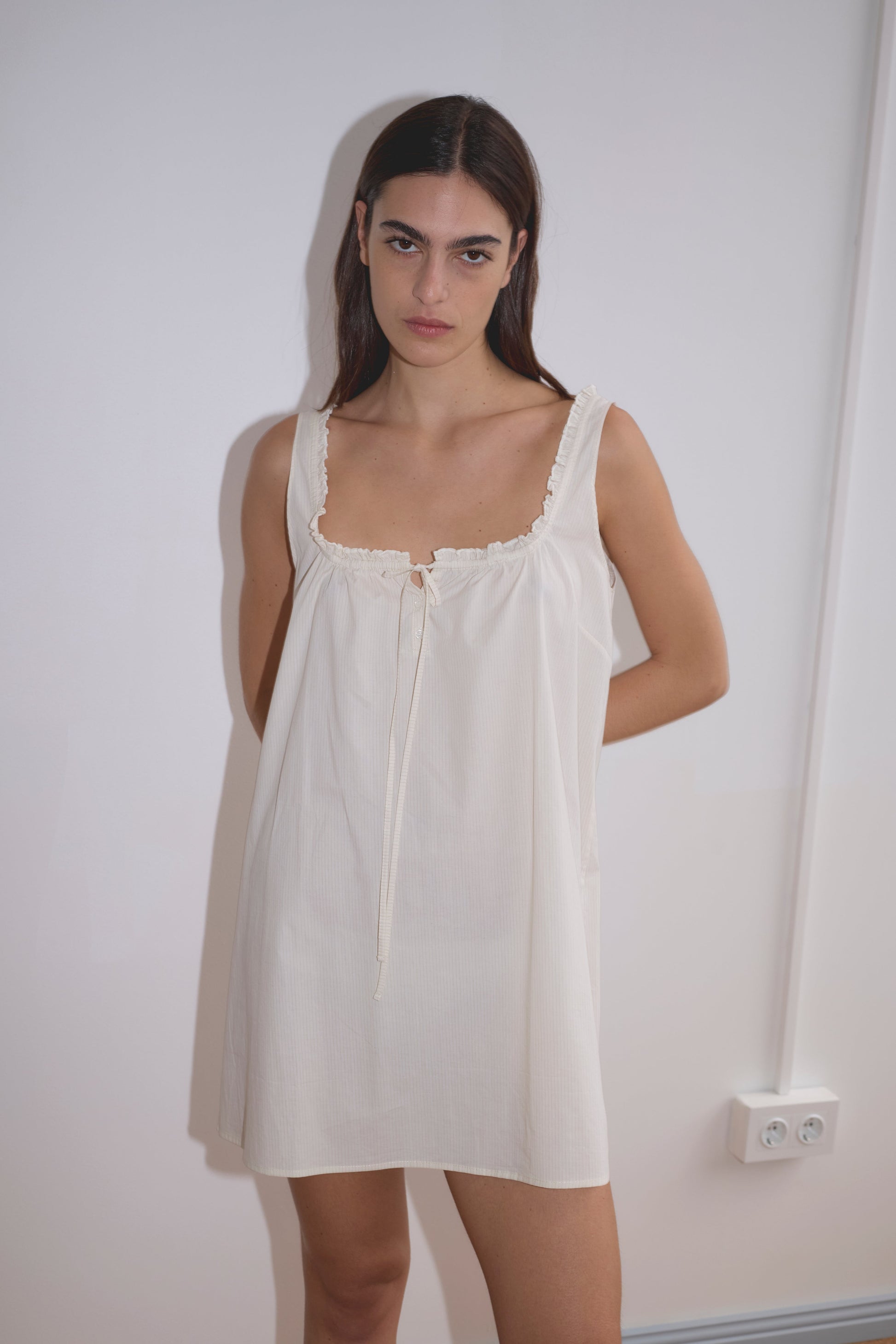 A woman with long dark hair wears the Button Frill Mini Dress in Stone Stripe by Deiji Studios, standing against a plain white wall and looking at the camera with a neutral expression. Two electrical outlets appear near her side.