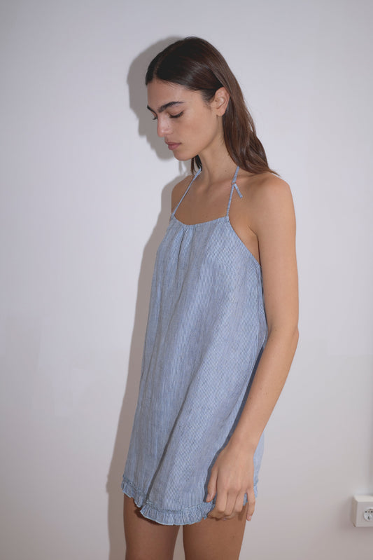 A woman with long brown hair wears the Flounce Halter Dress - Blue Stripe by Deiji Studios, made from organic French linen, standing against a plain white wall and looking down with a neutral expression.