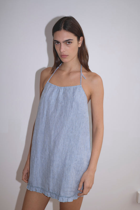 A woman with long dark hair wears the Flounce Halter Dress - Blue Stripe and stands against a plain white wall, looking at the camera with a neutral expression.