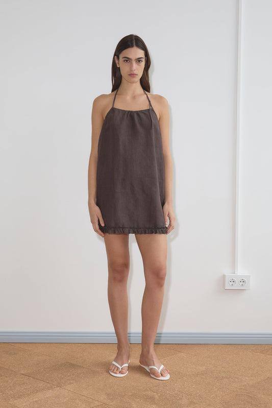 A woman stands indoors against a plain white wall, wearing the Flounce Halter Dress - Bark, a sleeveless dark brown French linen mini with a halter neck, paired with white sandals. Her arms are relaxed by her sides; a light brown floor and outlet are visible.