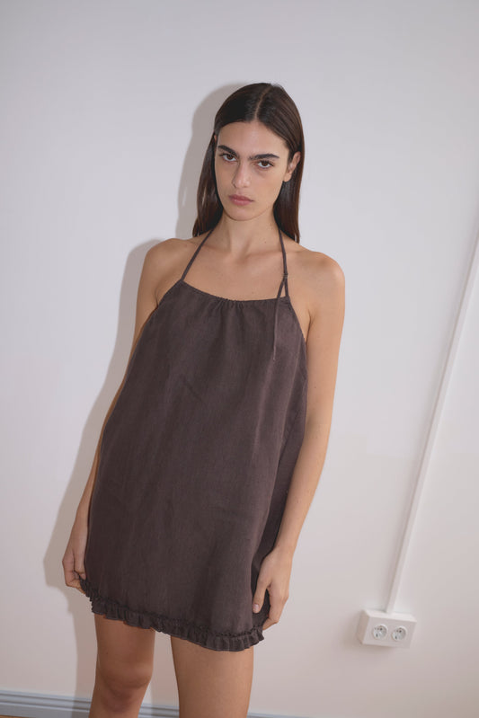 A woman with straight brown hair stands against a white wall, wearing the Flounce Halter Dress - Bark. The sleeveless dress features a halter neckline, loose fit, and ruffled hem. She looks at the camera with a neutral expression.