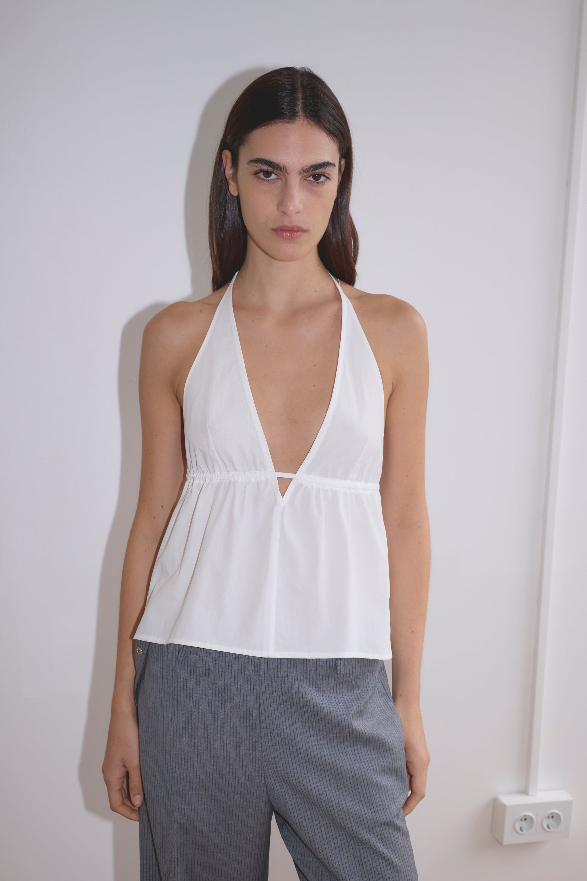 A person with long dark hair wears the Button Halter, a white deep V-neck top, paired with loose gray pants, standing against a plain white wall.