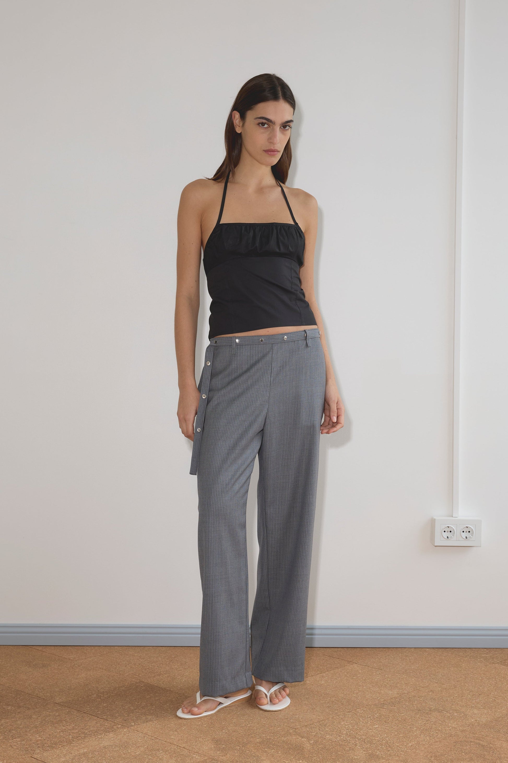 A woman with long brown hair and a neutral expression stands indoors against a plain wall, wearing the Deiji Studios Strap Top - Black and gray wide-leg side-button pants in organic cotton, paired with white sandals.