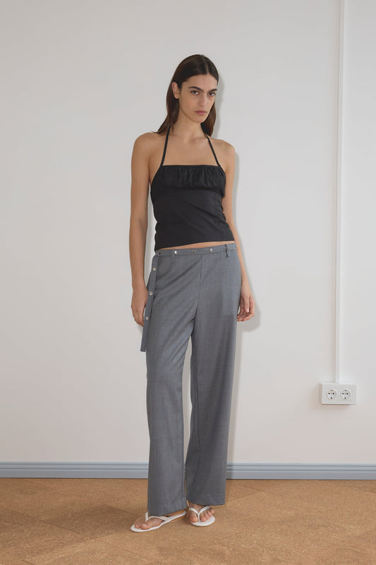 A woman with long brown hair and a neutral expression stands indoors against a plain wall, wearing the Deiji Studios Strap Top - Black and gray wide-leg side-button pants in organic cotton, paired with white sandals.