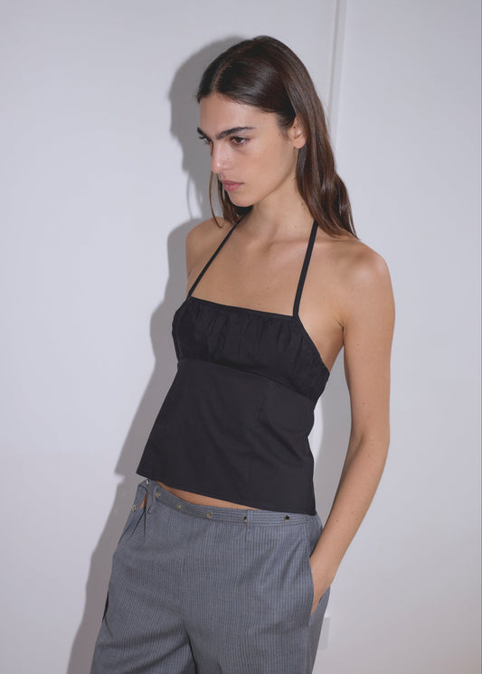 A woman with long brown hair stands against a plain white wall, wearing the Strap Top - Black by Deiji Studios and gray pinstripe pants, hands in her pockets and looking slightly downward.