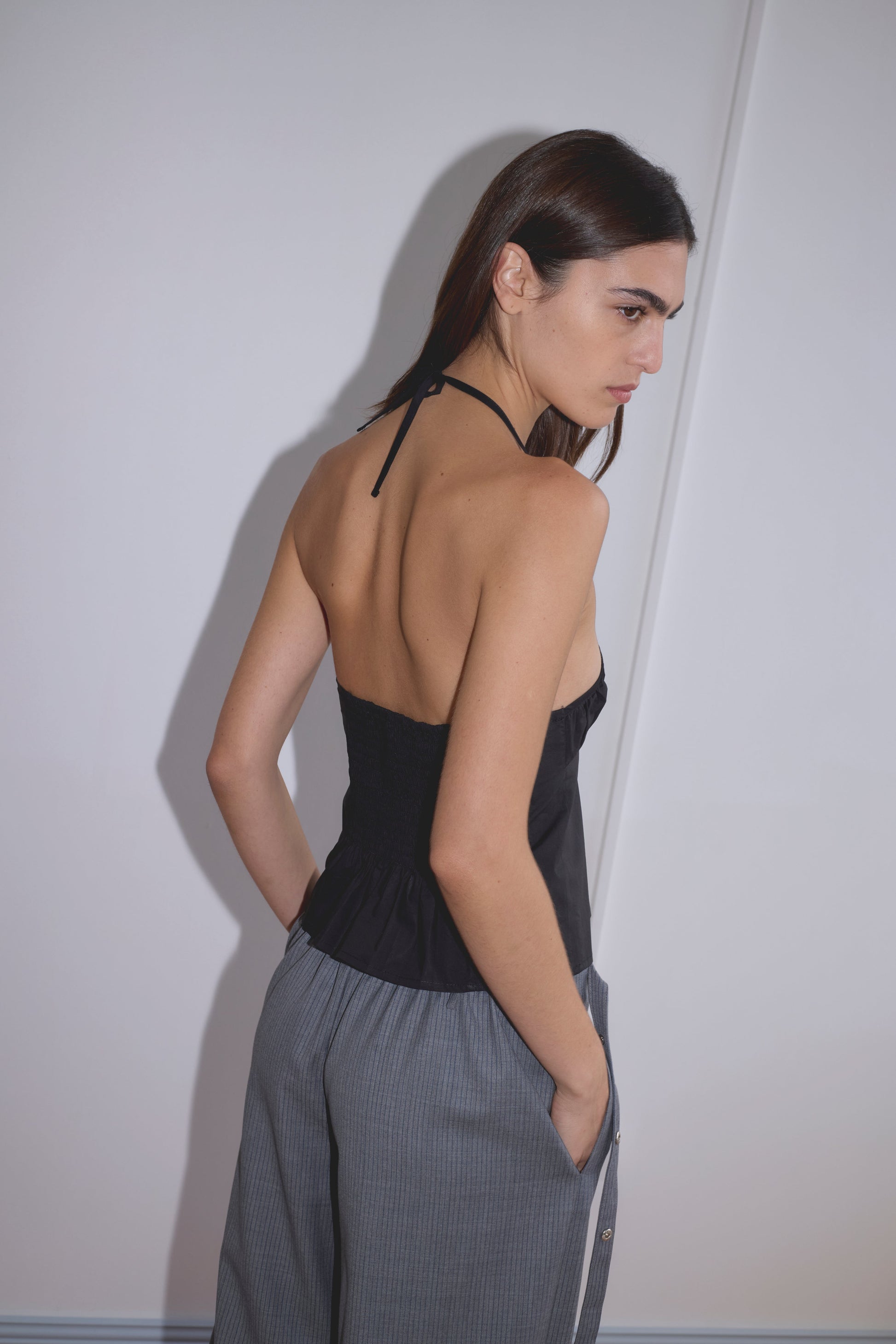 A woman with long brown hair stands sideways, looking over her shoulder. She wears the Strap Top - Black by Deiji Studios, featuring an open back, paired with gray high-waisted pants, her hands in the pockets against a plain white wall.