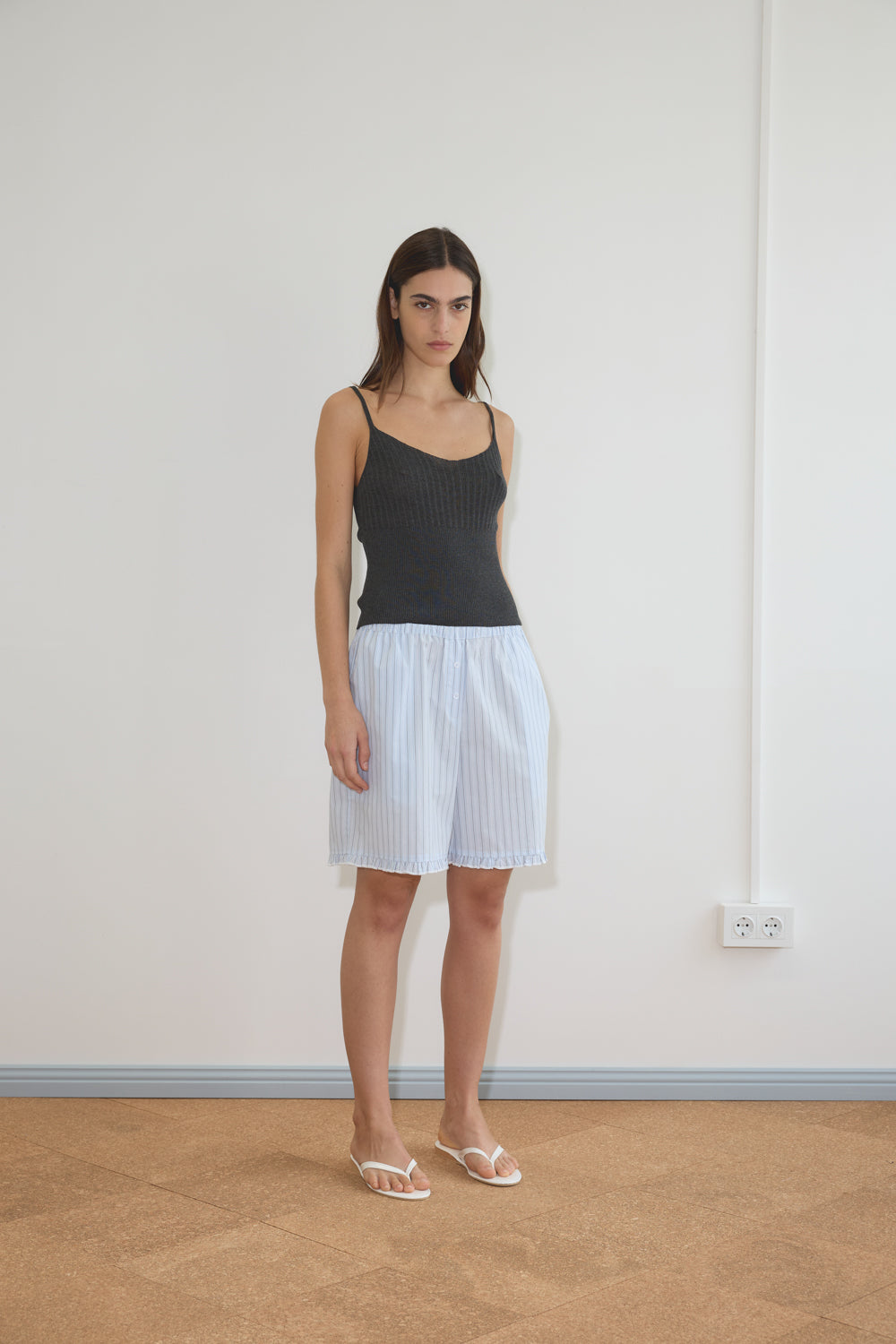 A woman with long brown hair wears the Ribbed Tank - Dark Charcoal by Deiji Studios, paired with a light blue and white striped skirt and white sandals, standing indoors on a brown floor near a white wall with an electrical outlet.