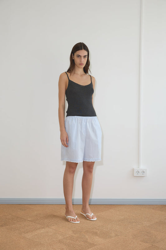 A woman with long brown hair wears the Ribbed Tank - Dark Charcoal by Deiji Studios, paired with a light blue and white striped skirt and white sandals, standing indoors on a brown floor near a white wall with an electrical outlet.