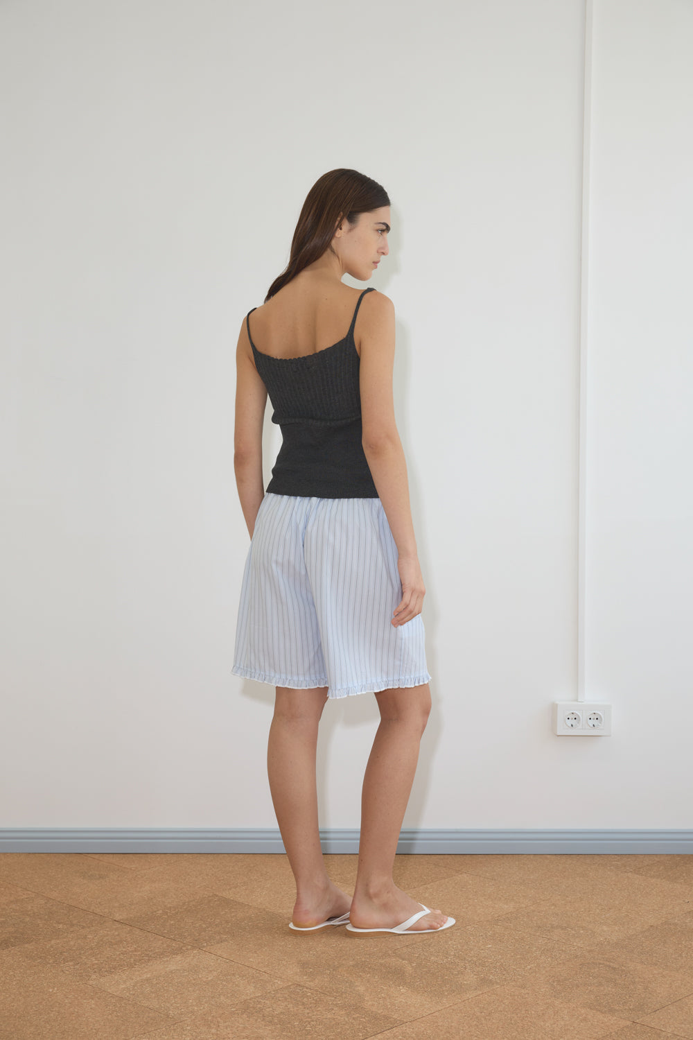 A woman with straight brown hair wears Deiji Studios' Ribbed Tank in Dark Charcoal, light blue knee-length pleated shorts, and white sandals. She stands indoors facing a white wall, with her back and profile visible.