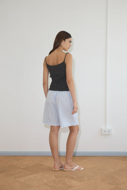 A woman with straight brown hair wears Deiji Studios' Ribbed Tank in Dark Charcoal, light blue knee-length pleated shorts, and white sandals. She stands indoors facing a white wall, with her back and profile visible.