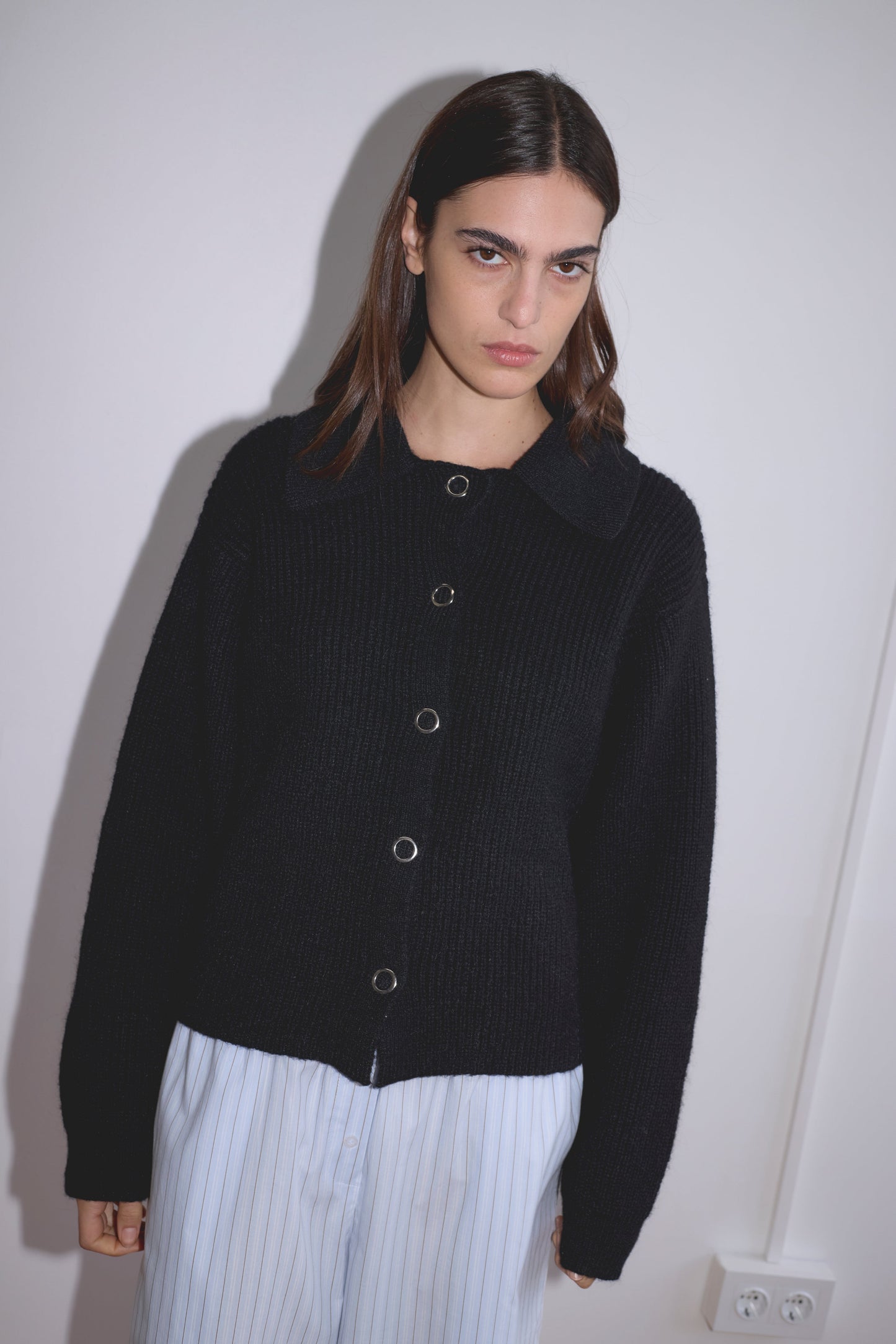 A woman with straight dark hair wears the Stud Cardi Knit in black, styled in an oversized fit, paired with light blue striped pants, standing against a white wall and looking at the camera with a neutral expression.