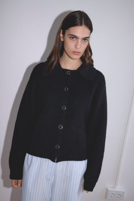 A woman with straight dark hair wears the Stud Cardi Knit in black, styled in an oversized fit, paired with light blue striped pants, standing against a white wall and looking at the camera with a neutral expression.