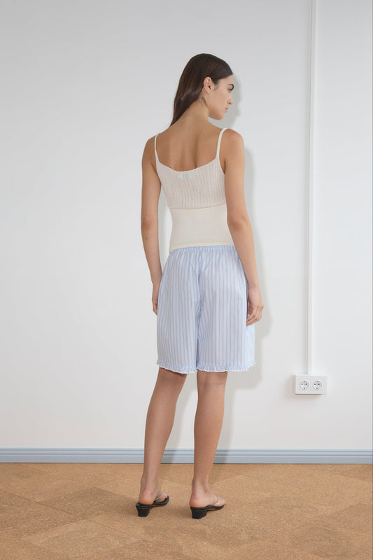 A woman stands indoors facing away, wearing the Ribbed Tank - Off White by Deiji Studios with loose, striped light blue shorts. She is barefoot on a tan floor near a white wall with two electrical outlets by the baseboard.