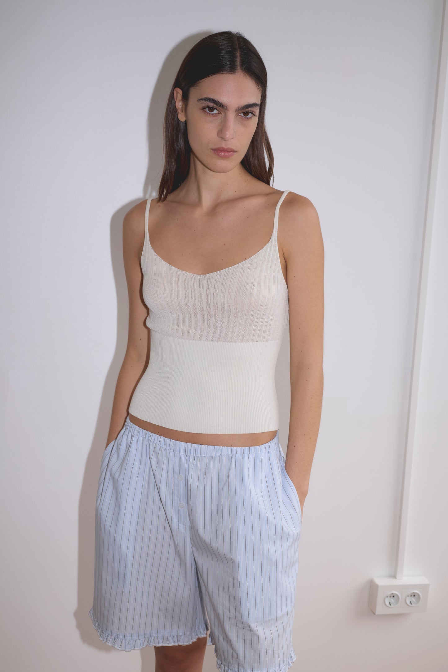 A woman with long dark hair stands against a white wall, hands in her pockets, wearing the Ribbed Tank in white and loose, light blue pinstriped shorts as she looks at the camera.