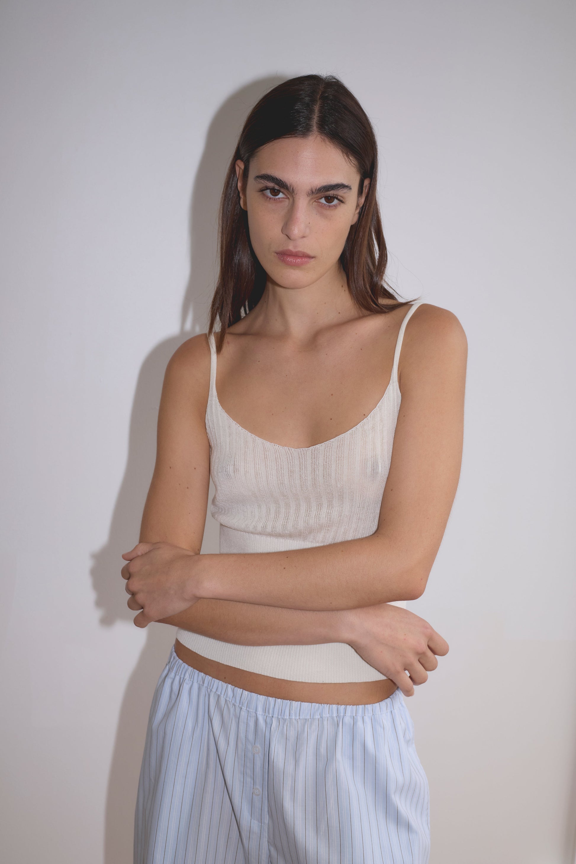 A woman with straight brown hair stands against a white wall, wearing Deiji Studios' Ribbed Tank - Off White and light blue striped pants, her arms folded loosely and her expression neutral.