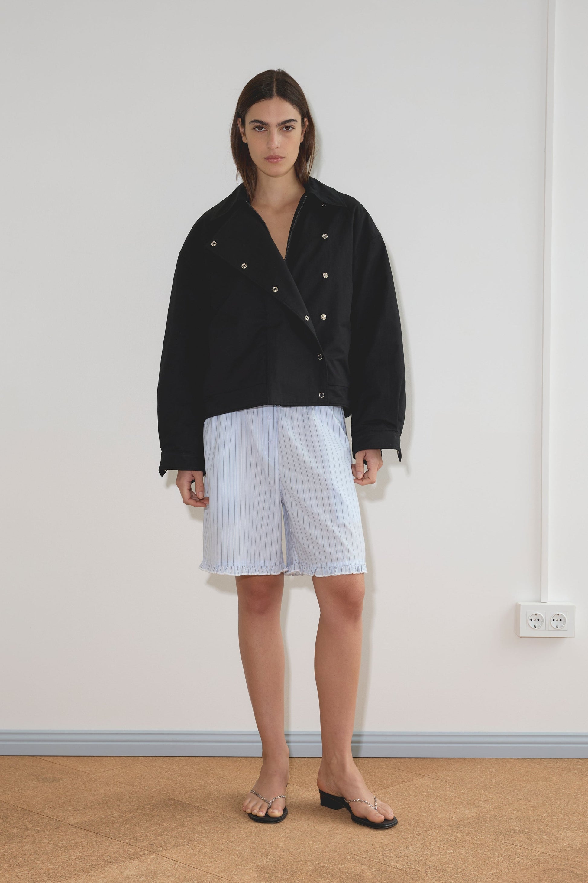 Indoors, a person poses against a plain white wall wearing the Lapel Jacket - Black, paired with light blue striped knee-length shorts and black heeled sandals. Their straight brown hair and neutral expression highlight the minimalist vibe.