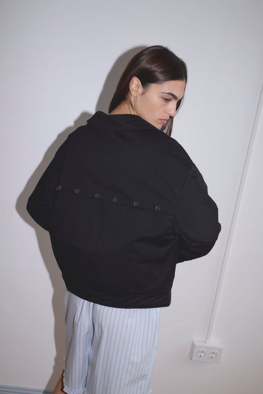 A person with long brown hair faces away, turning slightly, wearing a Deiji Studios Lapel Jacket - Black and light blue striped shorts in a minimalist room with white walls and a visible power outlet.