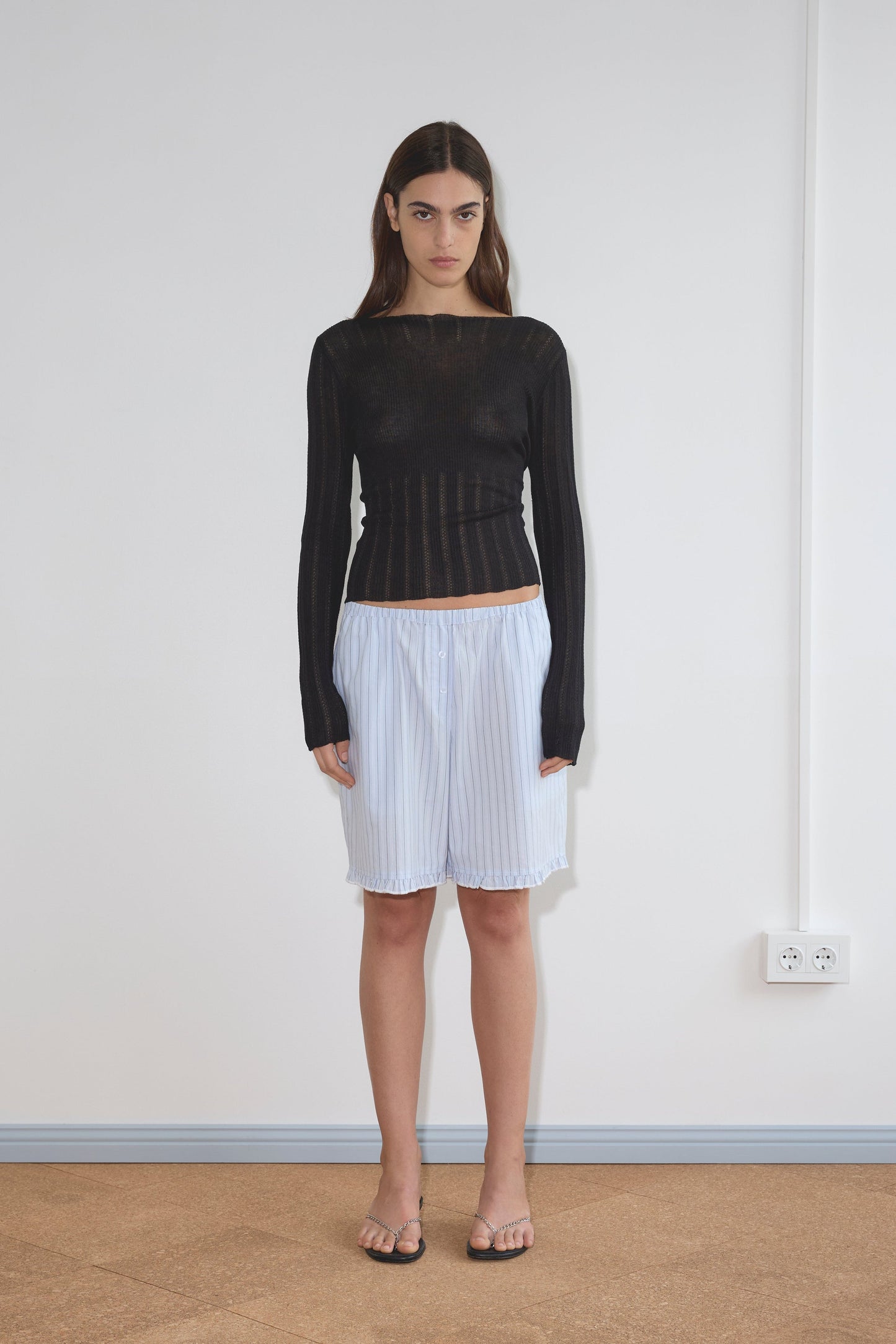 A woman with long brown hair and a neutral expression stands indoors against a white wall, wearing a sheer black long-sleeve top, the Flounce Short - Mineral Stripe with a relaxed fit, and sandals.