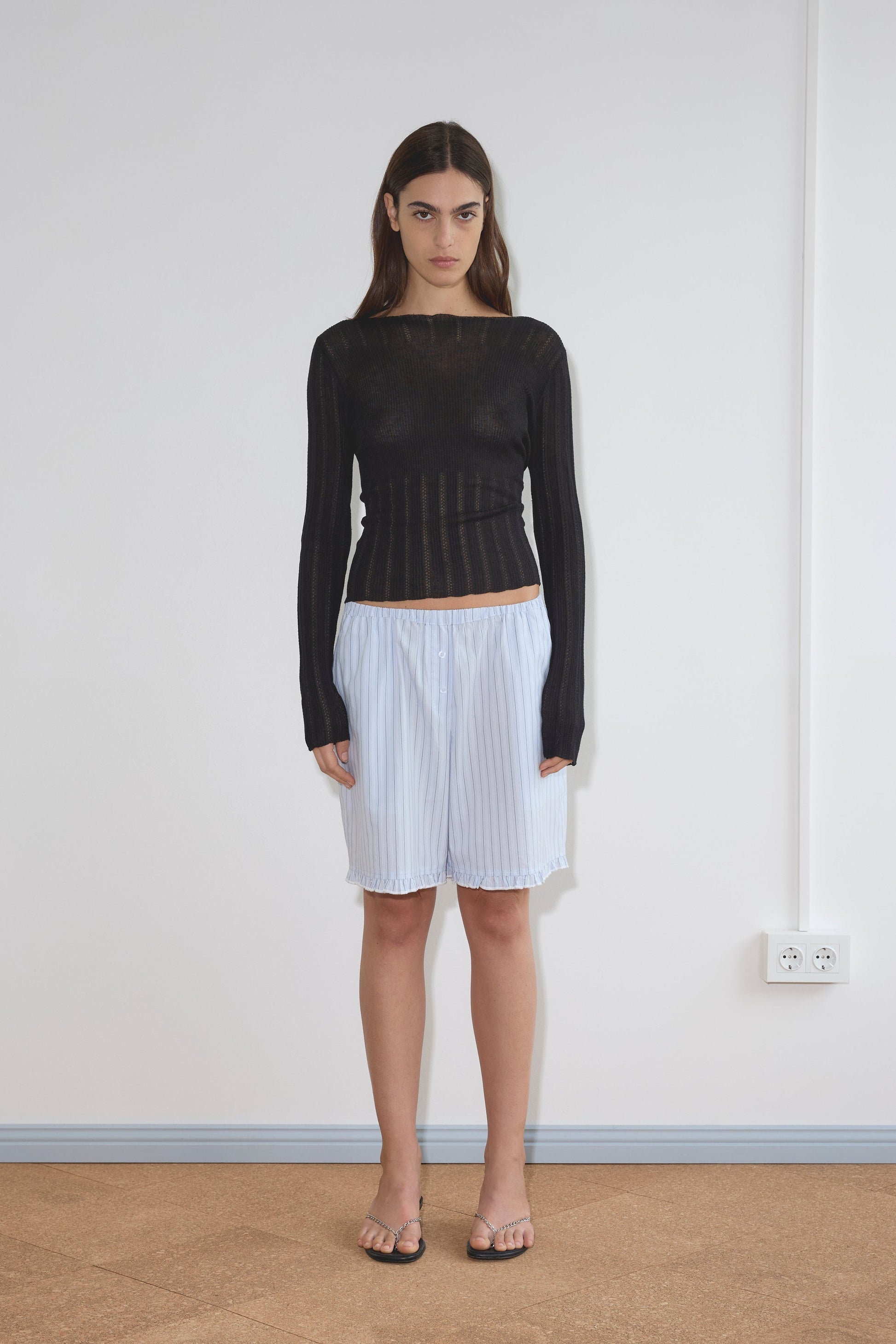 A woman with long brown hair and a neutral expression stands indoors against a white wall, wearing a sheer black long-sleeve top, the Flounce Short - Mineral Stripe with a relaxed fit, and sandals.