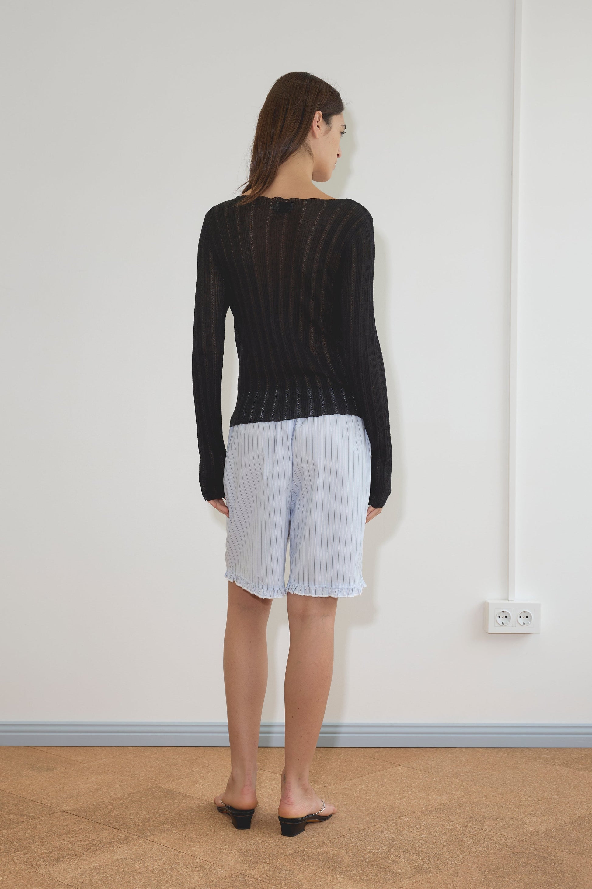 A woman stands indoors facing a white wall, wearing the Flounce Short - Mineral Stripe with a sheer black long-sleeve top. Her hair is straight, and she is barefoot on a light brown floor.