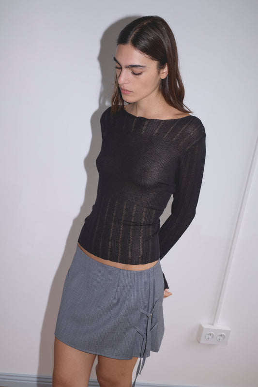 A woman with long brown hair stands against a white wall, wearing a sheer black long-sleeve top and the Tab Tie Skirt - Grey Blue Stripe. She looks down, and a power outlet is visible on the wall behind her.