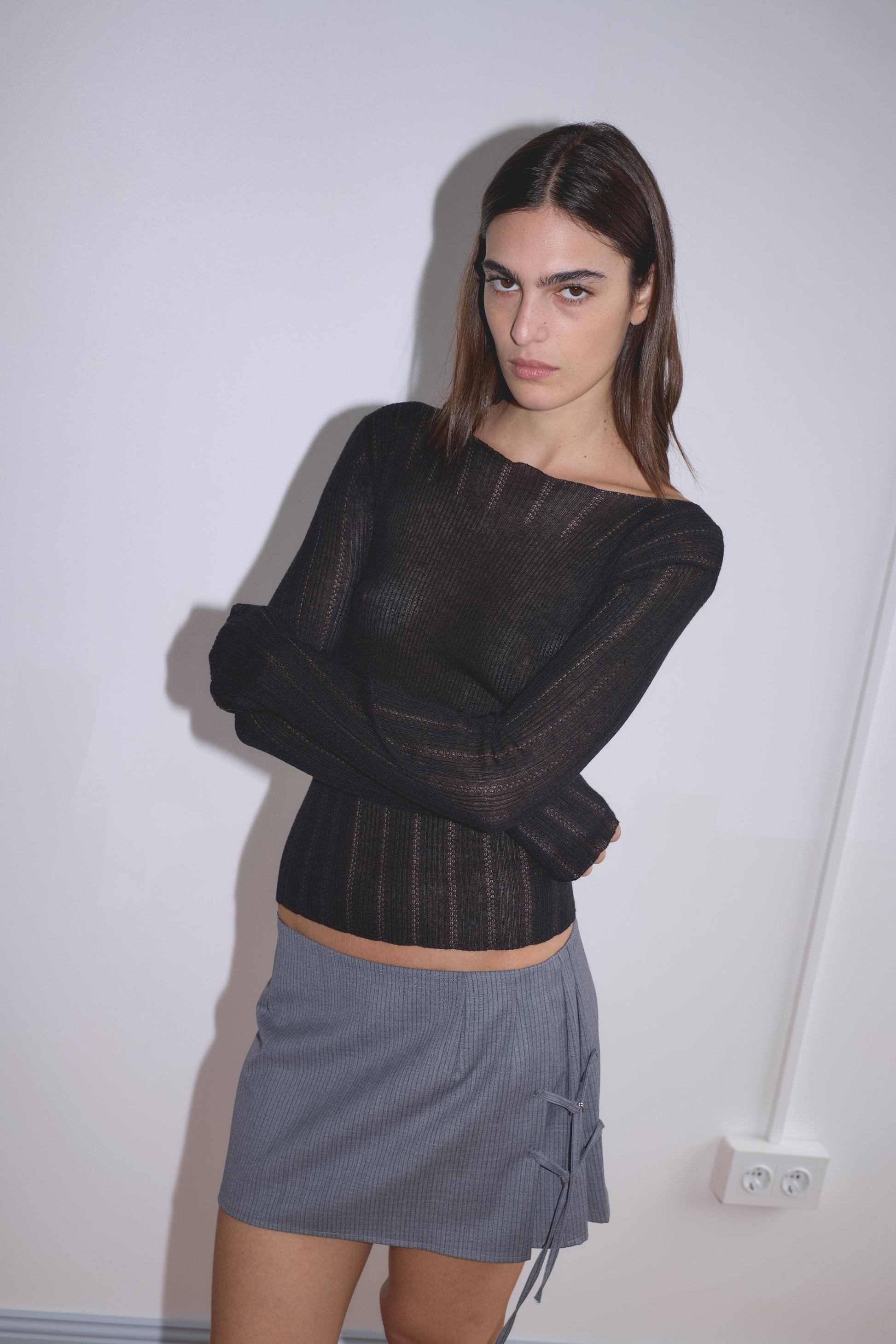 A woman with straight brown hair stands against a plain white wall, arms crossed. She wears a long-sleeved black sheer top and the Tab Tie Skirt - Grey Blue Stripe, reminiscent of Deiji Studios. An electrical outlet is visible beside her.