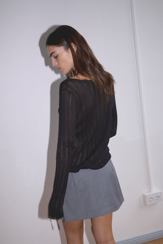 A woman with long brown hair stands sideways against a white wall, wearing a sheer black long-sleeve top and the Tab Tie Skirt - Grey Blue Stripe from Deiji Studios, looking down with a neutral expression.