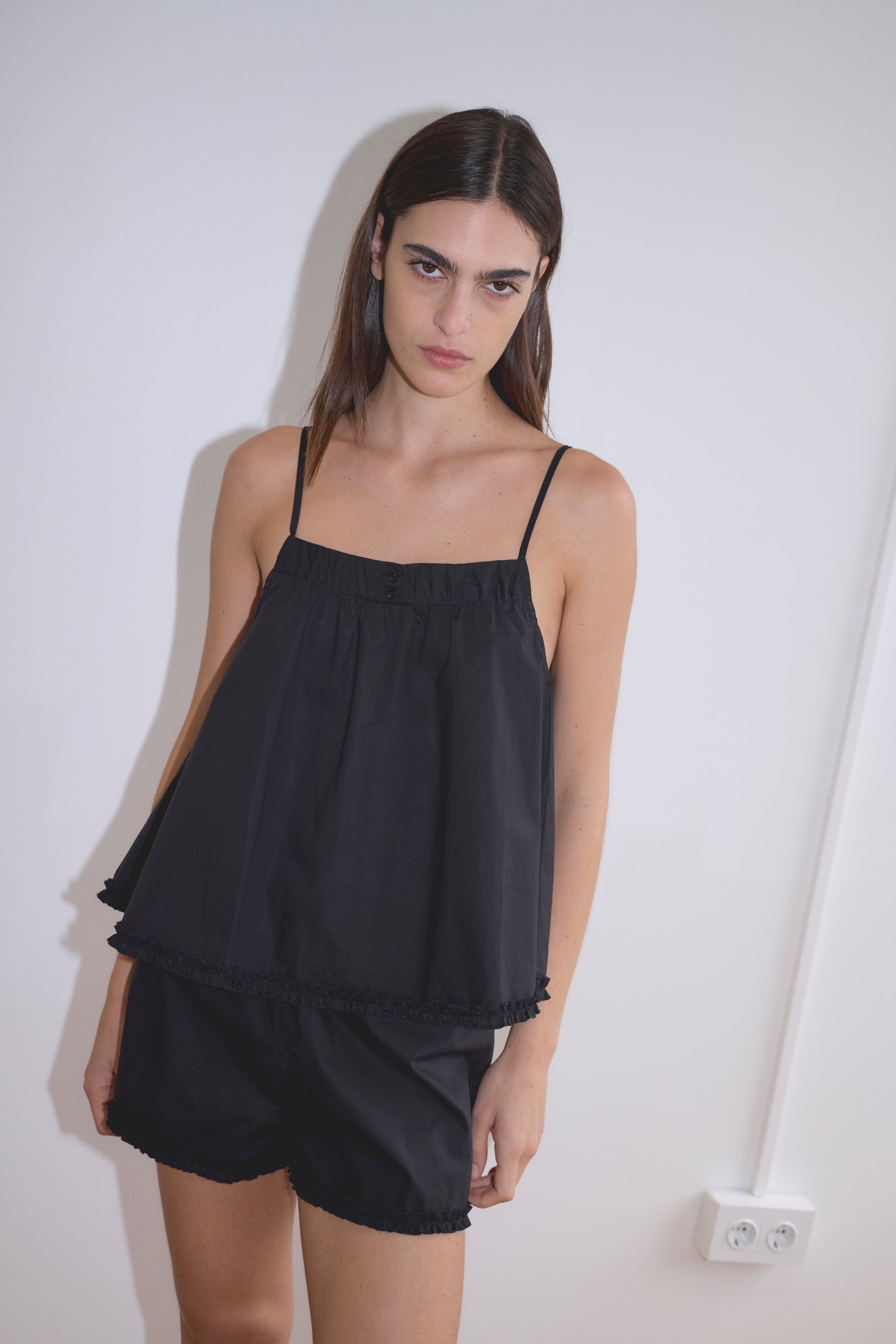 A woman with long dark hair stands against a white wall, wearing the Yoke Set - Black by Deiji Studios: a sleeveless organic cotton poplin top with thin straps and matching black shorts, her expression neutral and one hand resting by her side.