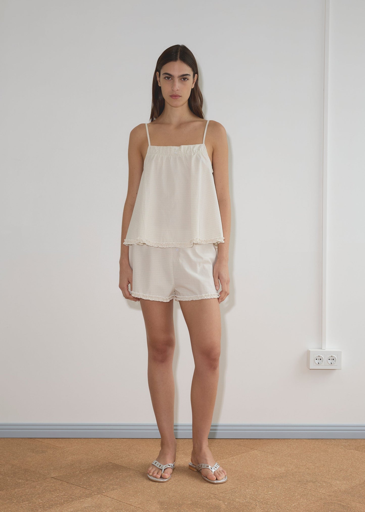 A woman with long brown hair and a neutral expression stands indoors against a white wall, wearing Deiji Studios’ Yoke Set - Bone Check, a loose organic cotton poplin babydoll top with matching shorts, paired with silver sandals.
