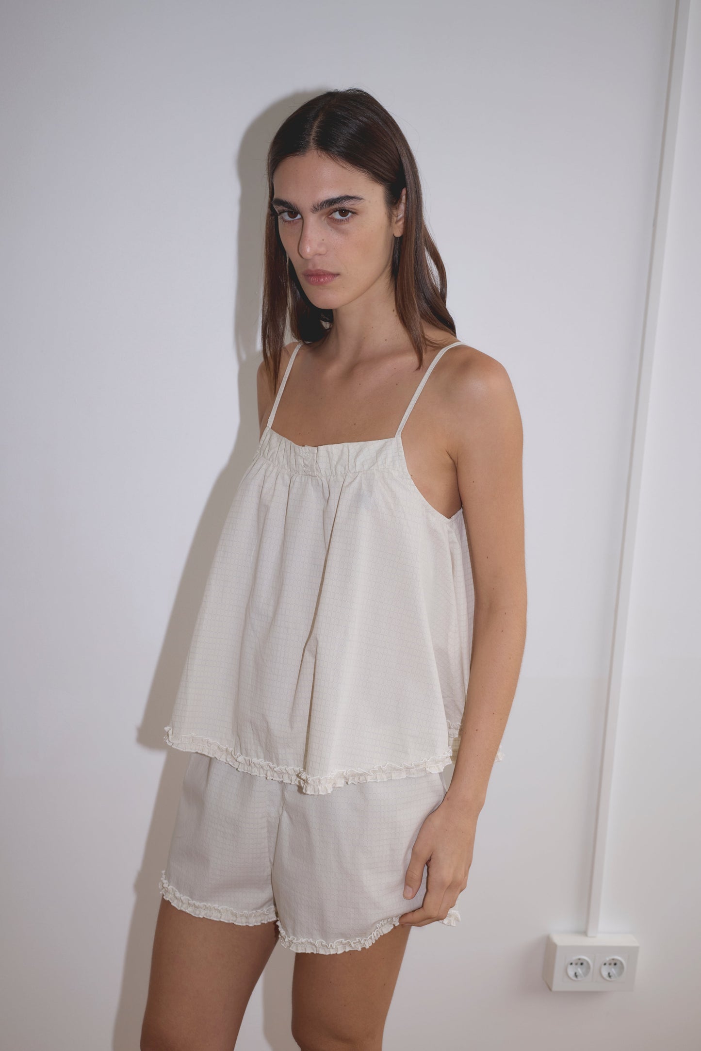 A woman with long dark hair stands against a white wall, wearing the Yoke Set - Bone Check from Deiji Studios, featuring a loose-fitting babydoll top and matching ruffled shorts in light organic cotton poplin. She poses with a neutral expression, hand on her hip.