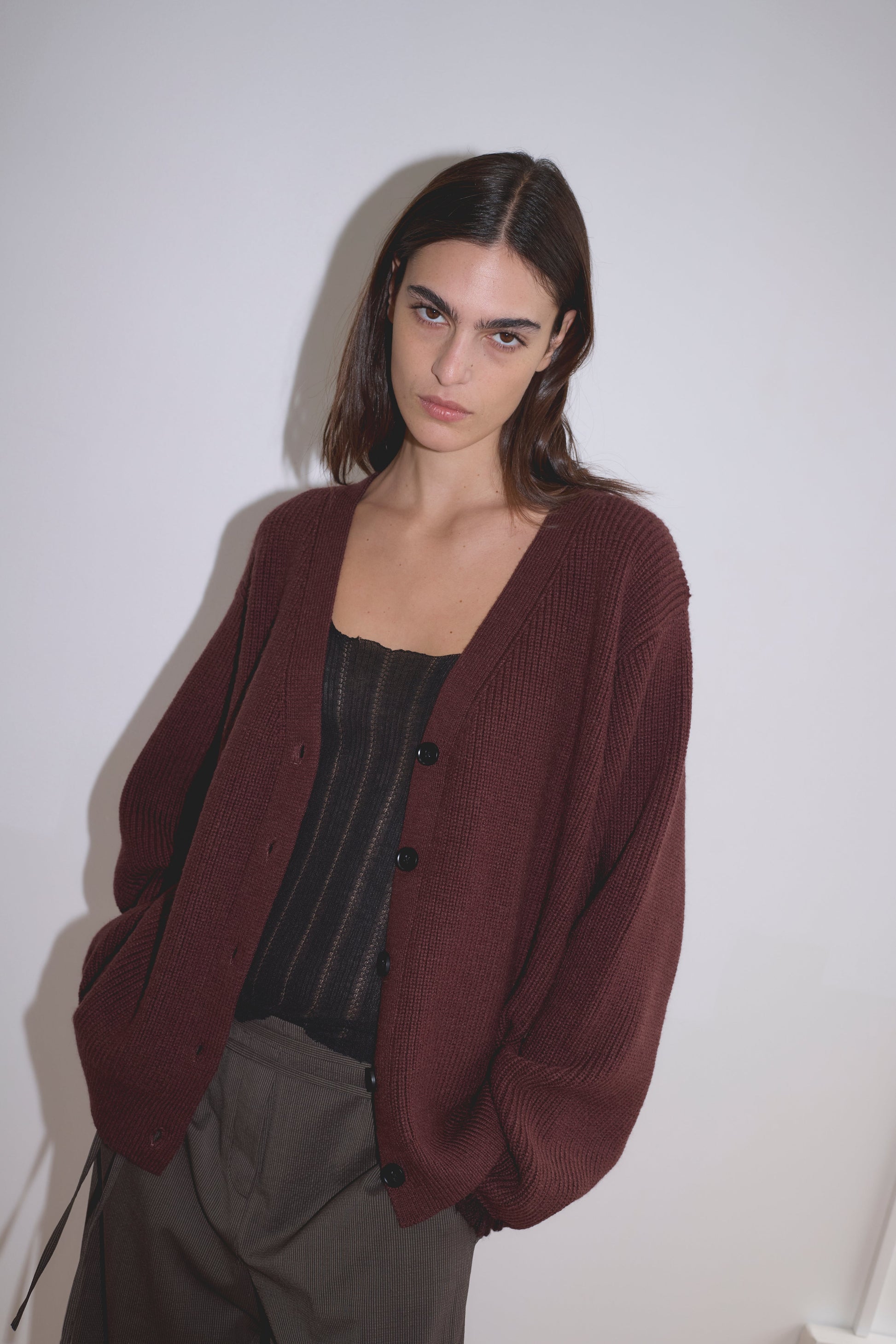 A person with straight brown hair wears the Flounce Cardigan - Burgundy by Deiji Studios over a black top and brown pants, standing against a plain white background with a neutral expression.