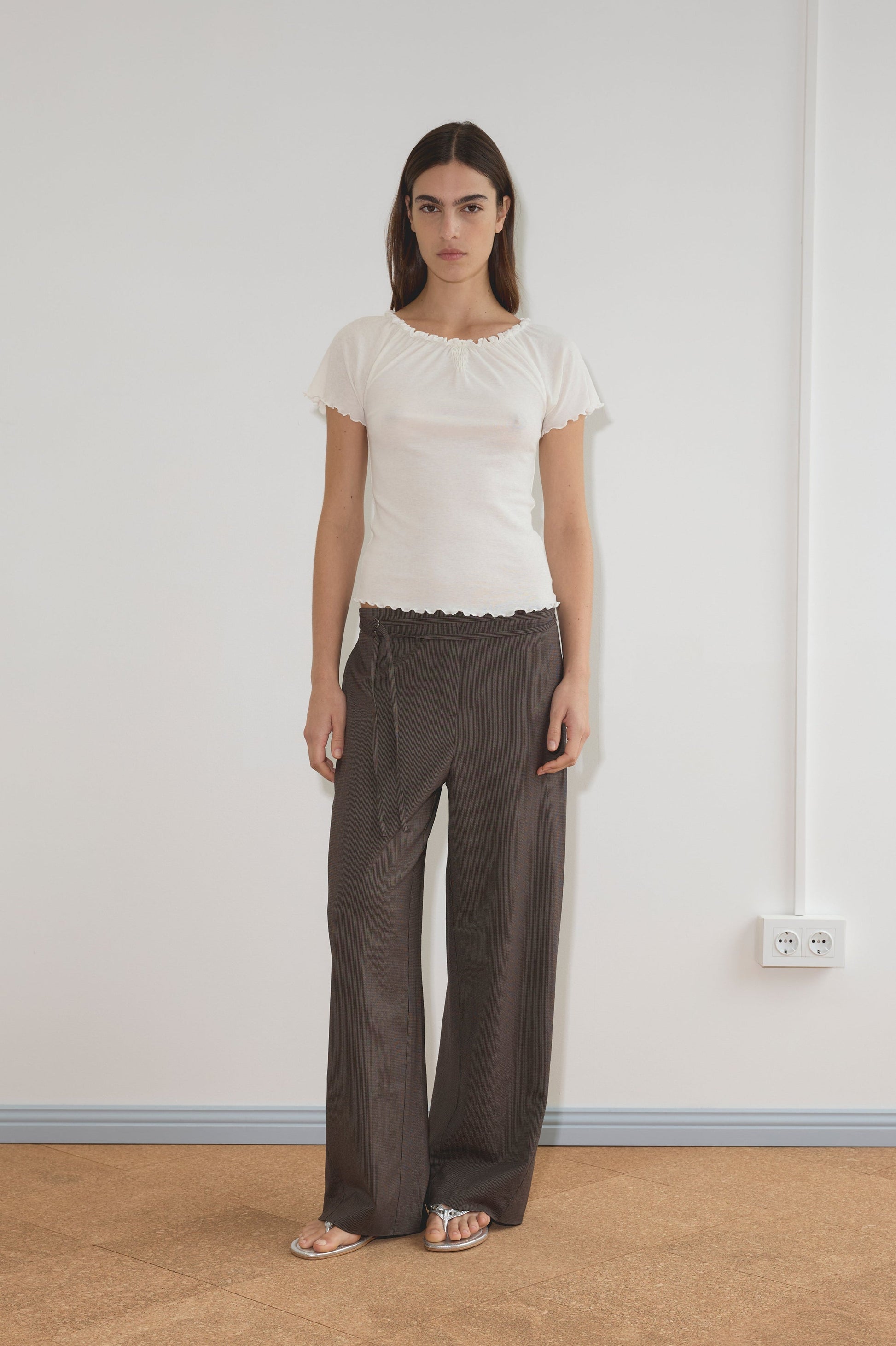 A person stands indoors against a plain white wall, wearing the Flounce Tee - Cream by Deiji Studios, loose brown trousers, and silver sandals. The floor is tan, with an electrical outlet visible on the right.