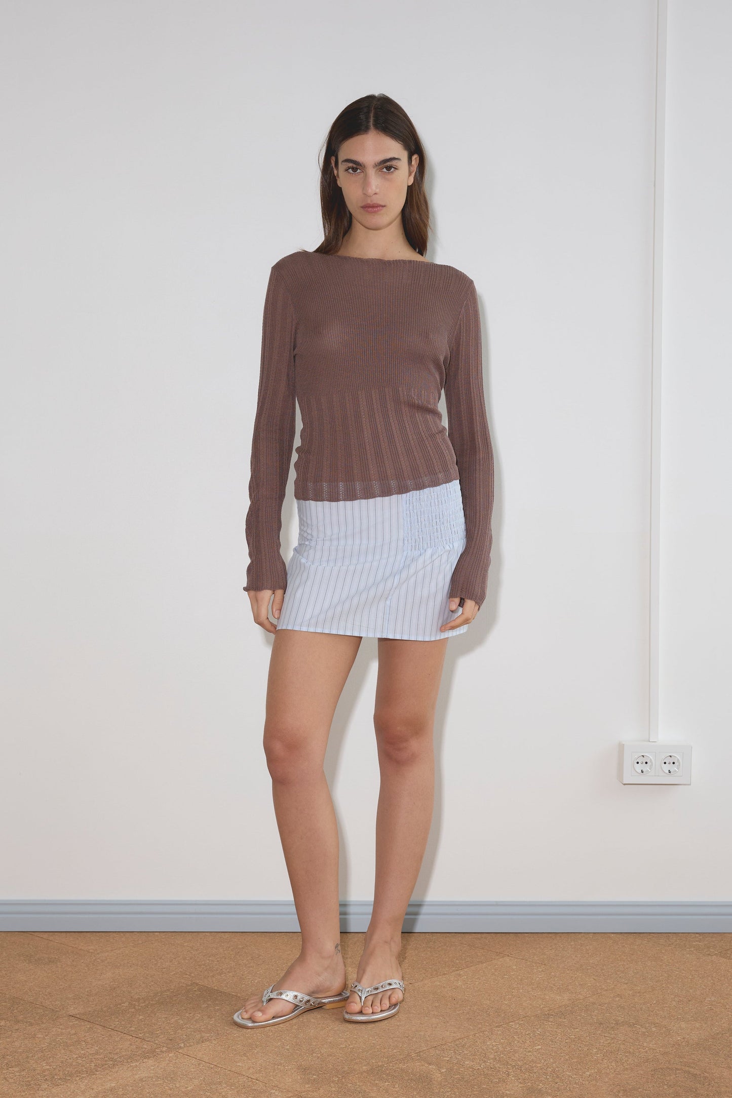 A woman with straight brown hair and a neutral expression stands indoors against a white wall, wearing a long-sleeved brown top, the Shirring Skirt - Mineral Stripe from Deiji Studios, made of organic cotton, and sandals.