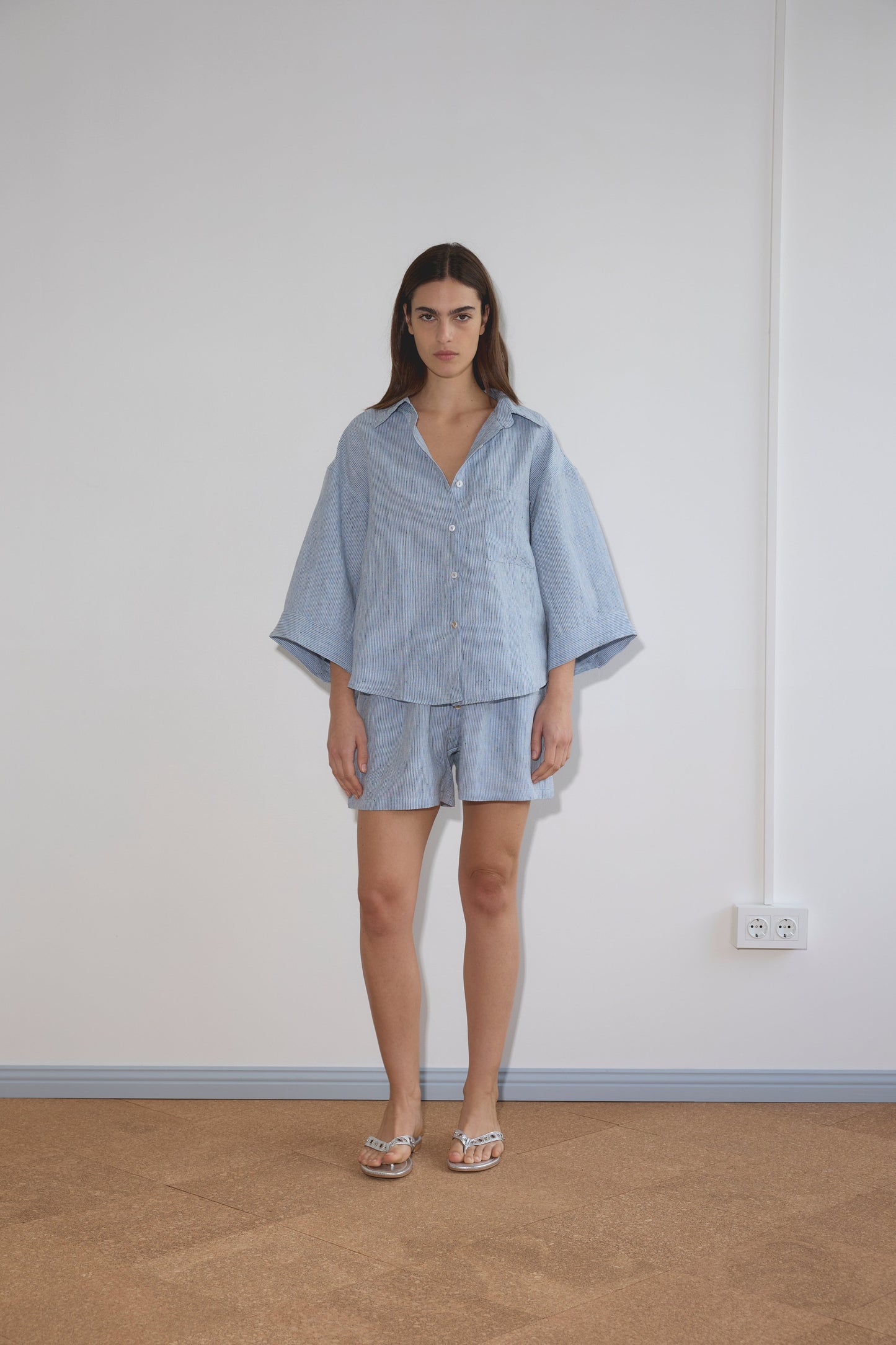 A woman stands indoors against a plain white wall, wearing the 03 Set - Blue Stripe: a loose-fitting blue and white striped shirt with wide sleeves and matching boxer shorts. She has long brown hair and silver sandals.