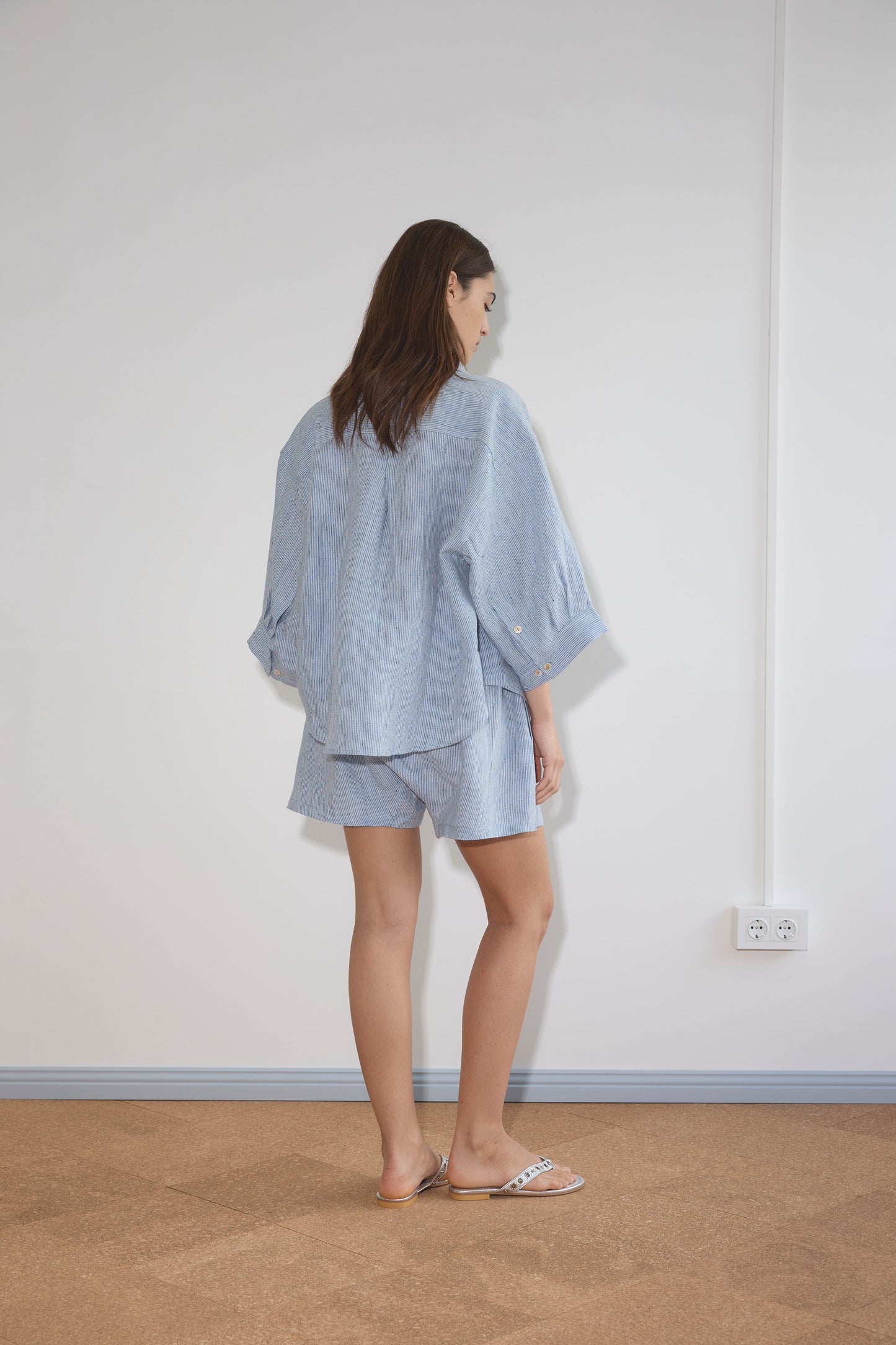 A woman with medium-length brown hair and white sandals stands indoors, facing away, wearing the 03 Set - Blue Stripe, consisting of a loose, light blue organic linen shirt and matching boxer shorts. White wall and power outlets are in the background.