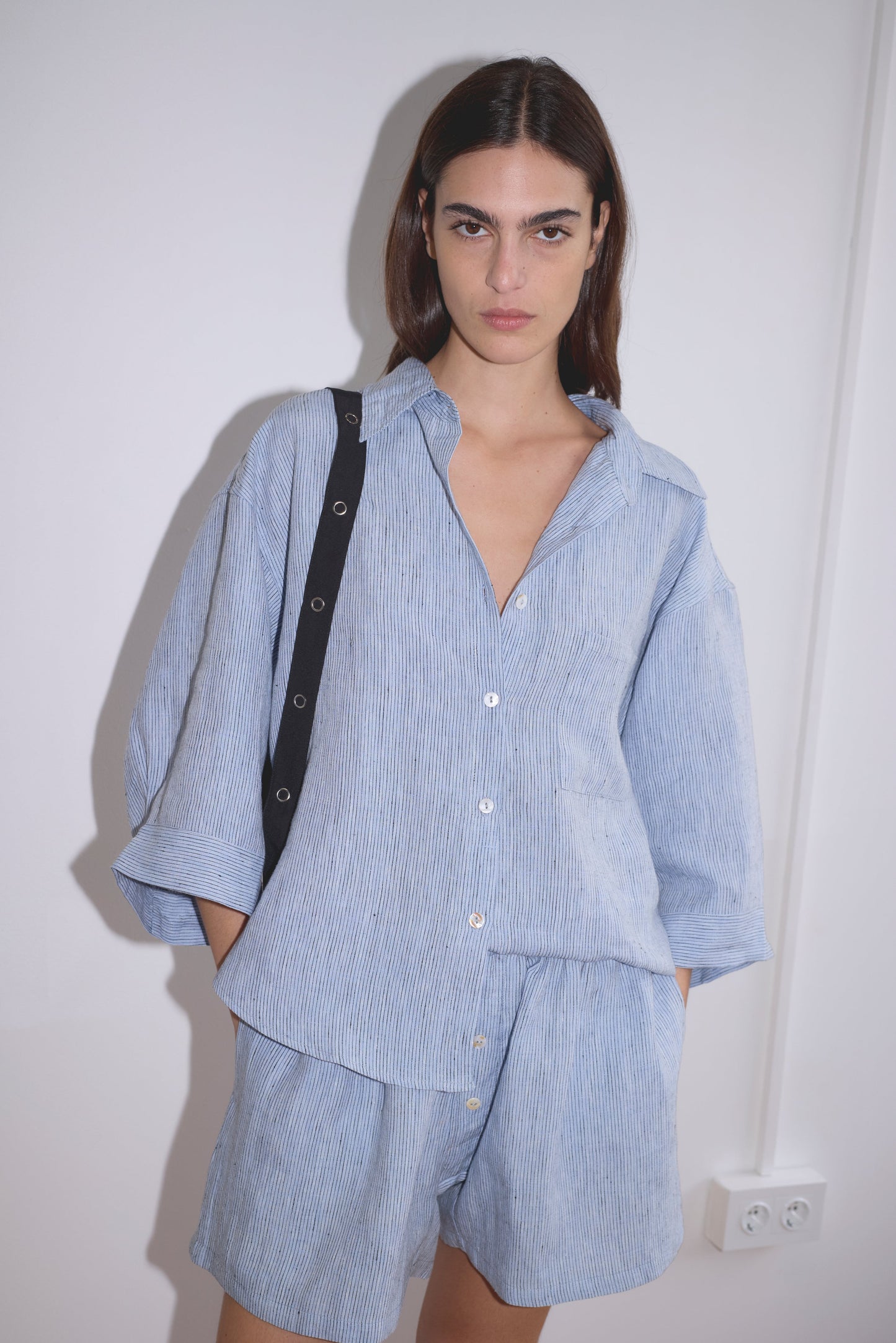 A woman with long dark hair wears the 03 Set - Blue Stripe by Deiji Studios—a loose linen shirt and matching boxer shorts—standing against a white wall, hands in pockets, looking at the camera.
