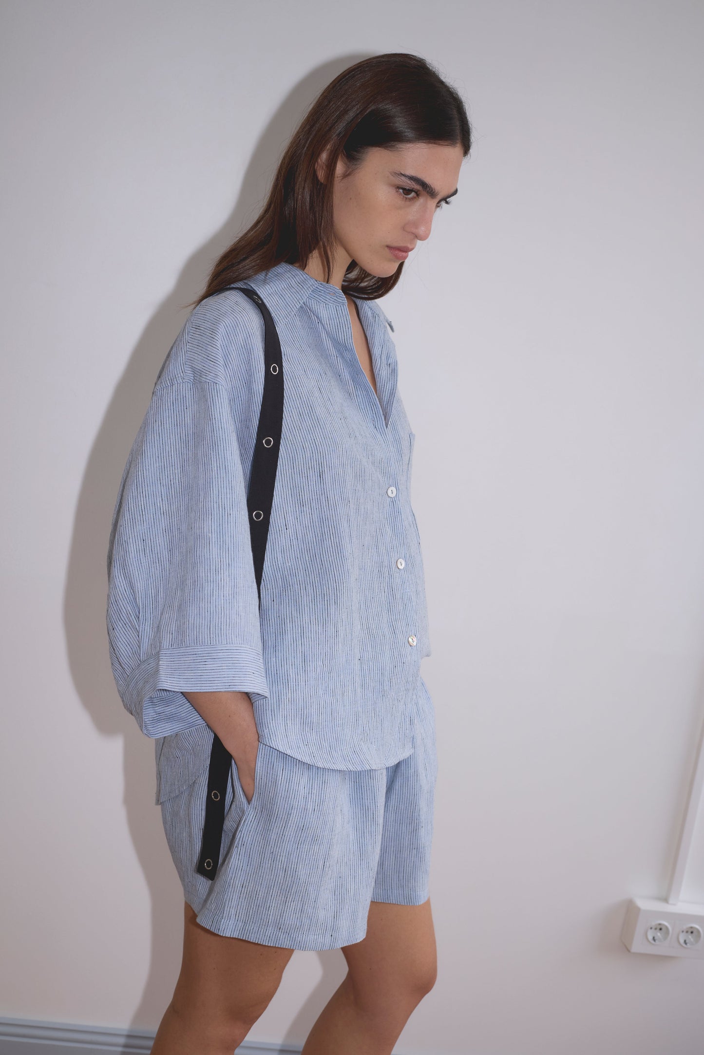 A woman with long brown hair wears the 03 Set - Blue Stripe: a loose-fitting organic linen shirt and matching boxer shorts, standing with hands in pockets against a plain, light-colored wall.