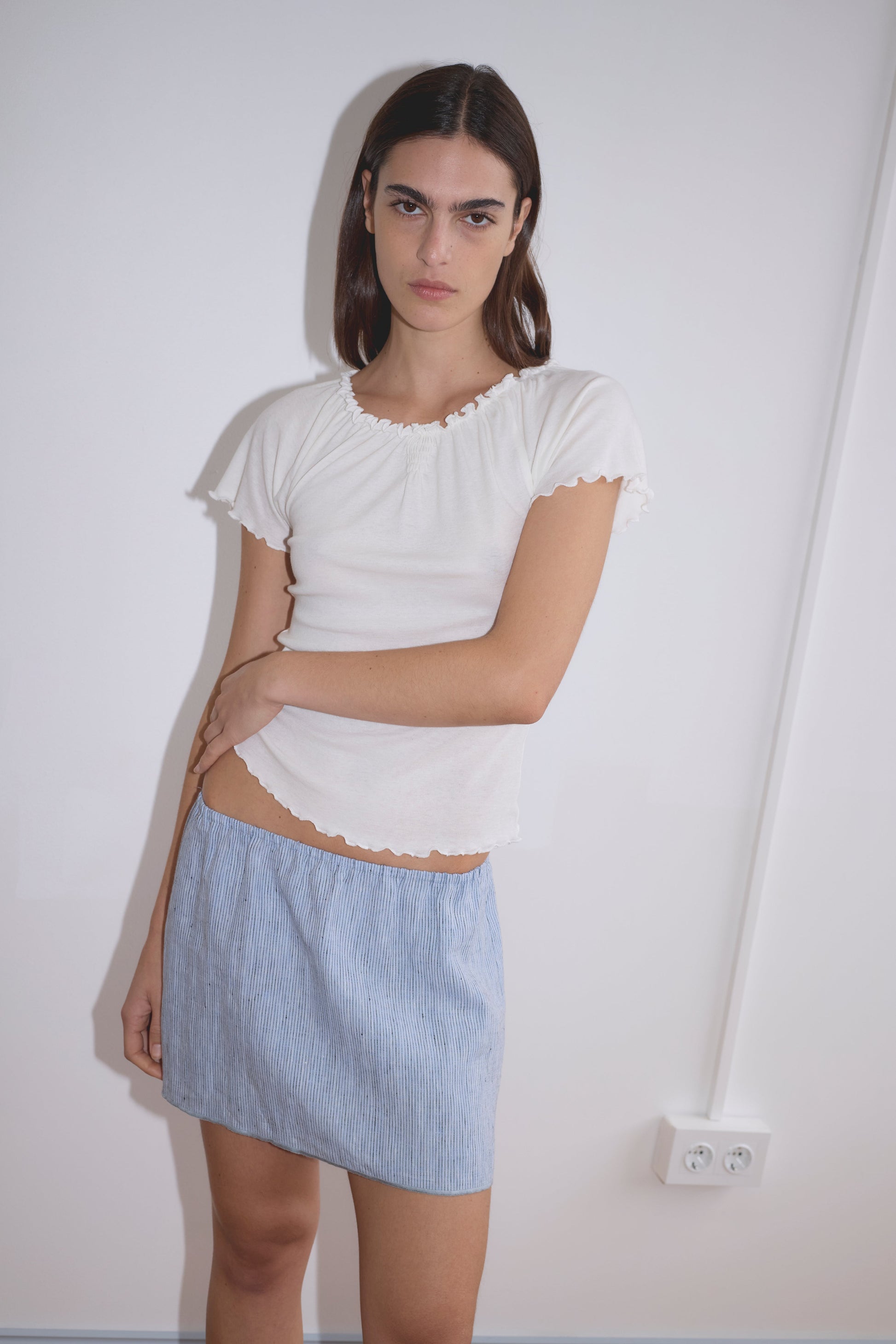 A person with straight brown hair wearing a white short-sleeved top and the Babylock Skirt - Blue Stripe stands against a white wall with a power outlet and visible cable in the background.