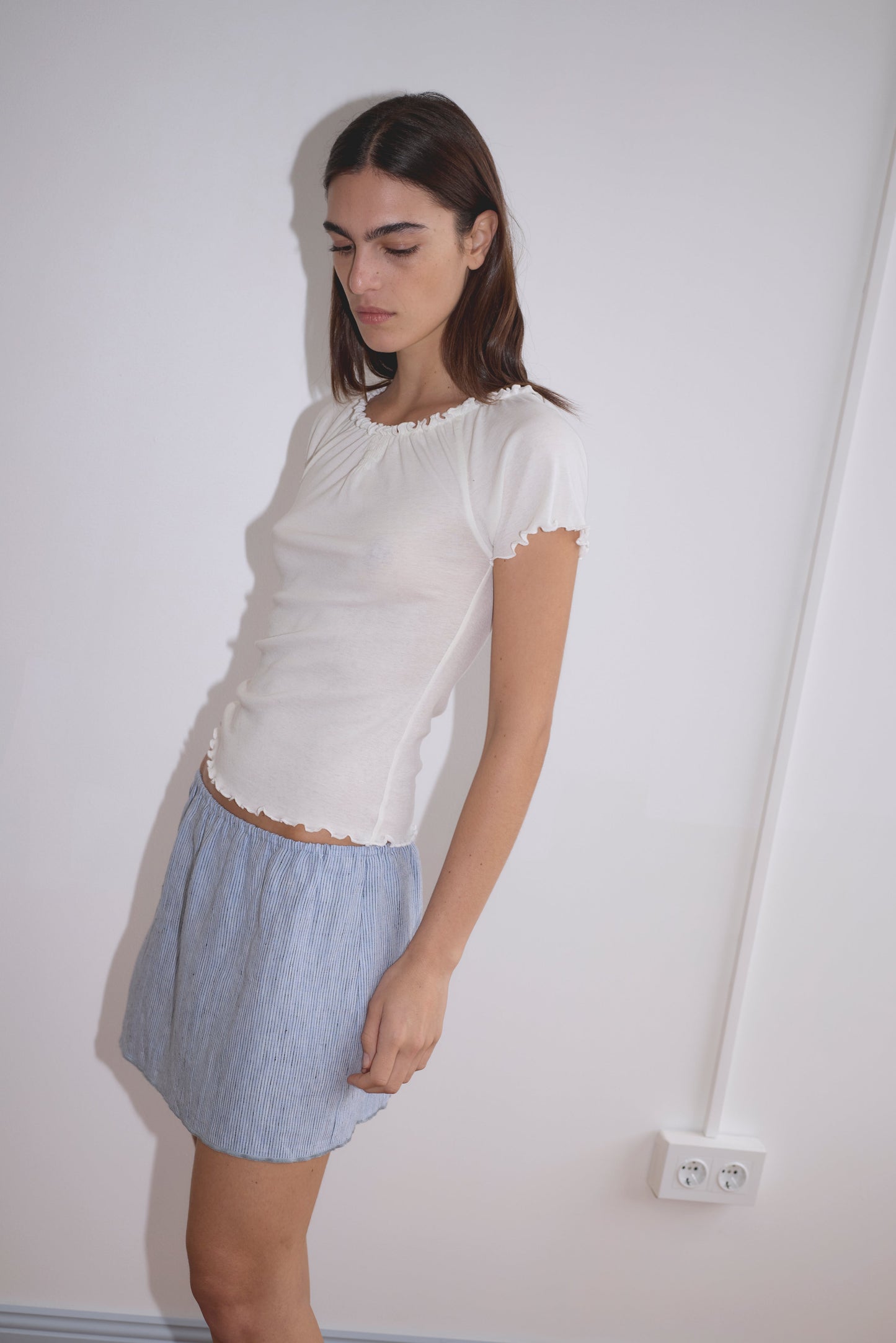 A woman with straight brown hair wears a fitted white short-sleeve top and the Babylock Skirt - Blue Stripe, standing against a plain white wall with a visible electrical outlet, looking down with a neutral expression.