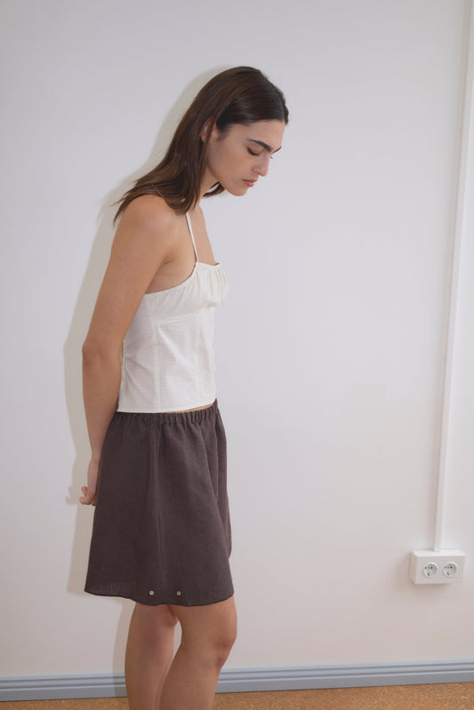 A woman with long brown hair stands sideways against a plain white wall, wearing a white sleeveless top and the Press Stud Short in dark brown, her hands behind her back as she looks down.