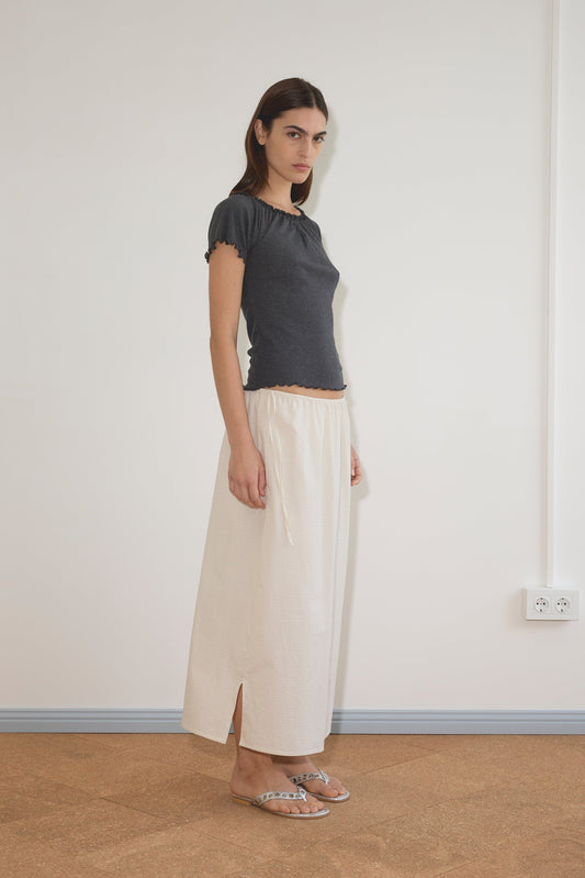 A woman stands indoors by a plain wall, wearing a dark short-sleeve top, flat sandals, and the cream-colored Mid Day Skirt with side slits. She faces sideways, looking at the camera with a neutral expression.
