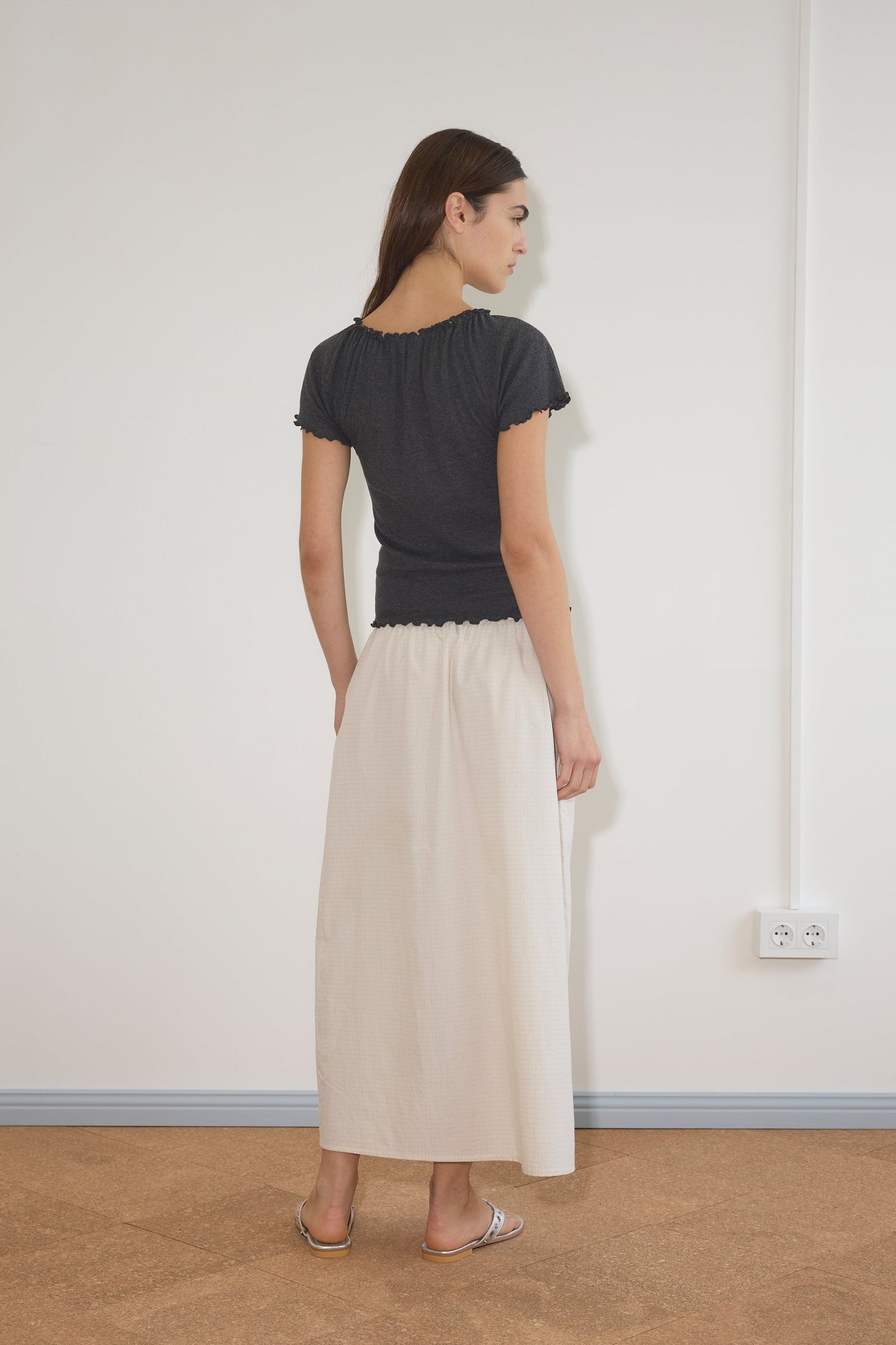 A woman stands indoors facing away, wearing a dark gray top and the Mid Day Skirt - Bone Check, a long cream organic cotton A-line skirt. She has straight brown hair and sandals, with a white wall and visible electrical outlet behind her.