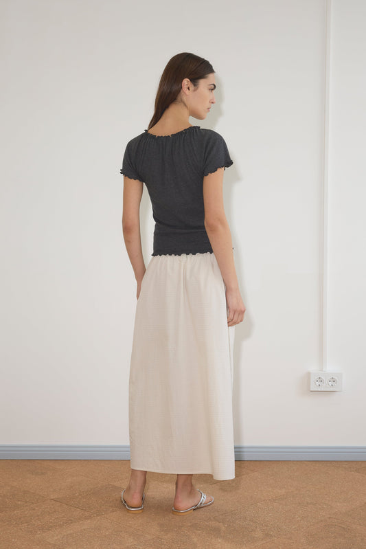 A woman stands indoors facing away, wearing a dark gray top and the Mid Day Skirt - Bone Check, a long cream organic cotton A-line skirt. She has straight brown hair and sandals, with a white wall and visible electrical outlet behind her.