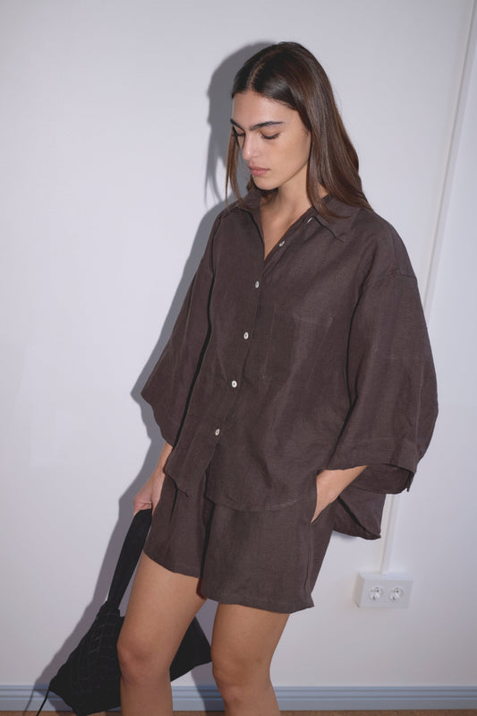 A woman stands against a white wall wearing the 03 Set - Bark: a loose, dark brown organic linen shirt and matching boxer-style shorts. One hand is in her pocket, the other holds a black bag as she looks down with a neutral expression.