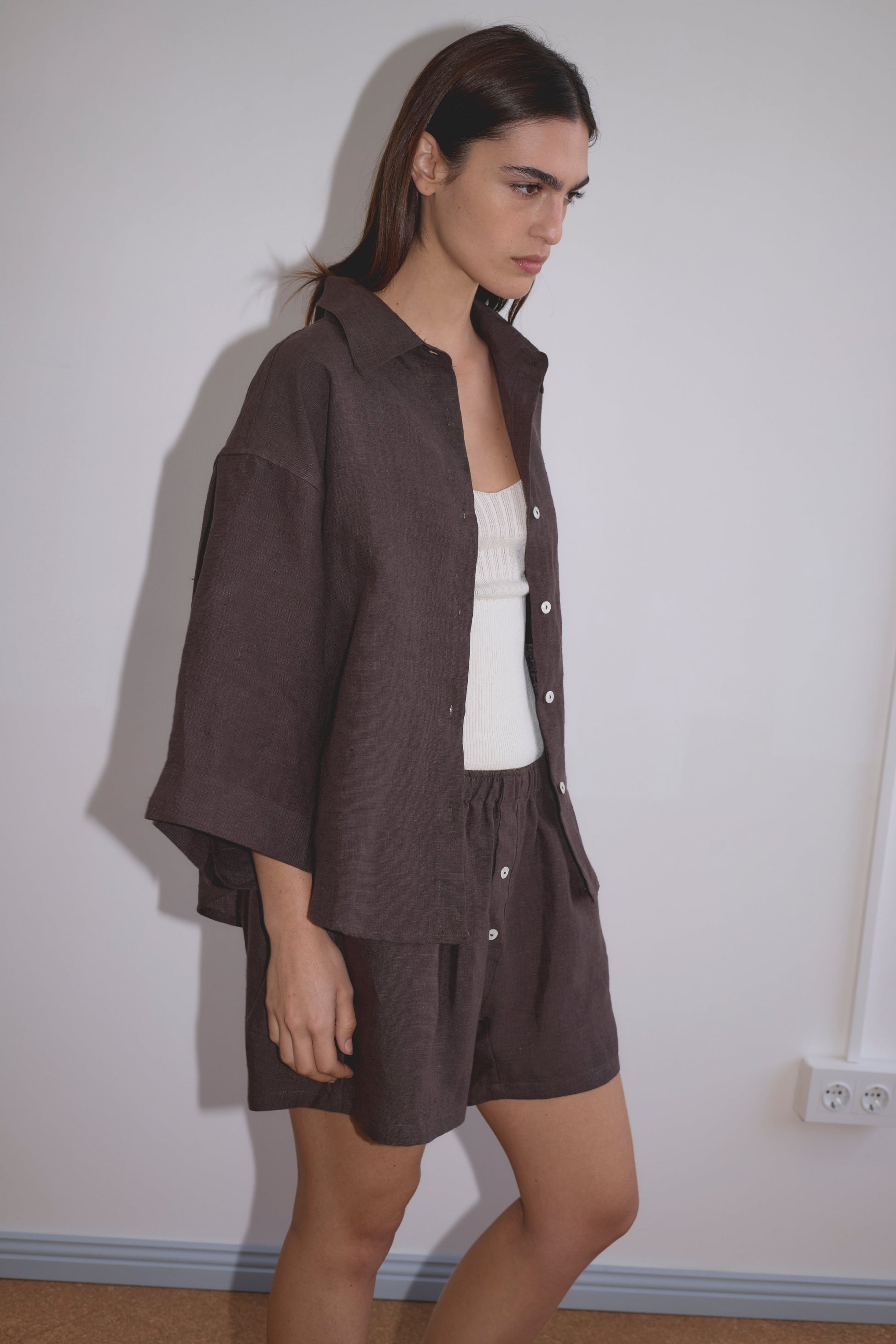 A woman with straight dark hair and a neutral expression stands against a pale wall, wearing the 03 Set - Bark: a loose dark brown organic linen shirt and matching boxer shorts layered over a white ribbed tank top.